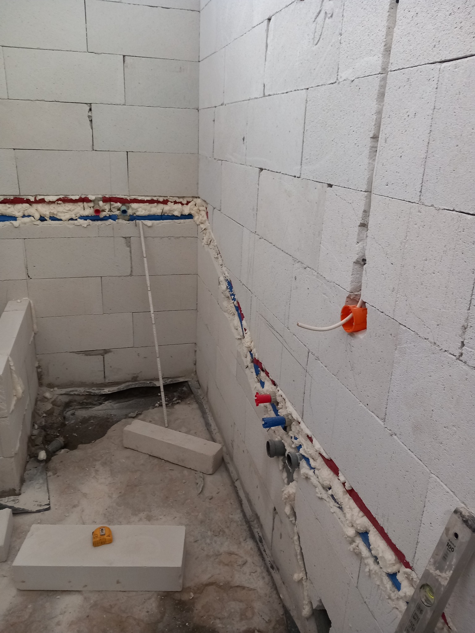 Surowa łazienka w trakcie prac: ściany z białych bloczków, instalacja wodna zabezpieczona pianką montażową, puszka elektryczna z przewodem, poziomica i miarka na podłodze.