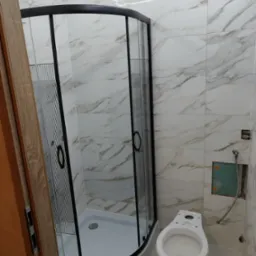 Nowa kabina prysznicowa z czarnymi profilami i brodzikiem, biała toaleta bez deski w łazience z marmurowymi płytkami, widoczne niedokończone prace przy instalacji.