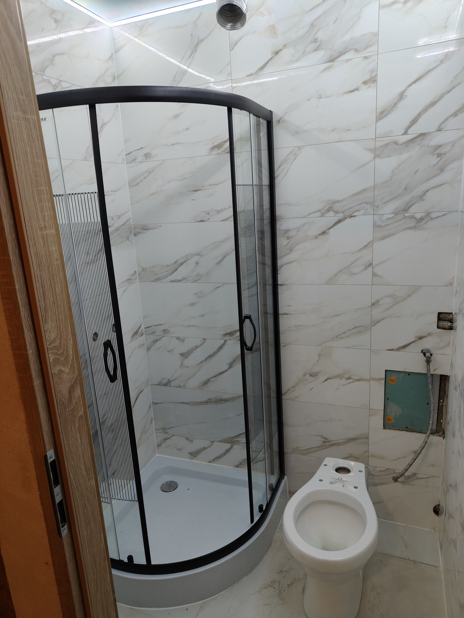 Nowa kabina prysznicowa z czarnymi profilami i brodzikiem, biała toaleta bez deski w łazience z marmurowymi płytkami, widoczne niedokończone prace przy instalacji.