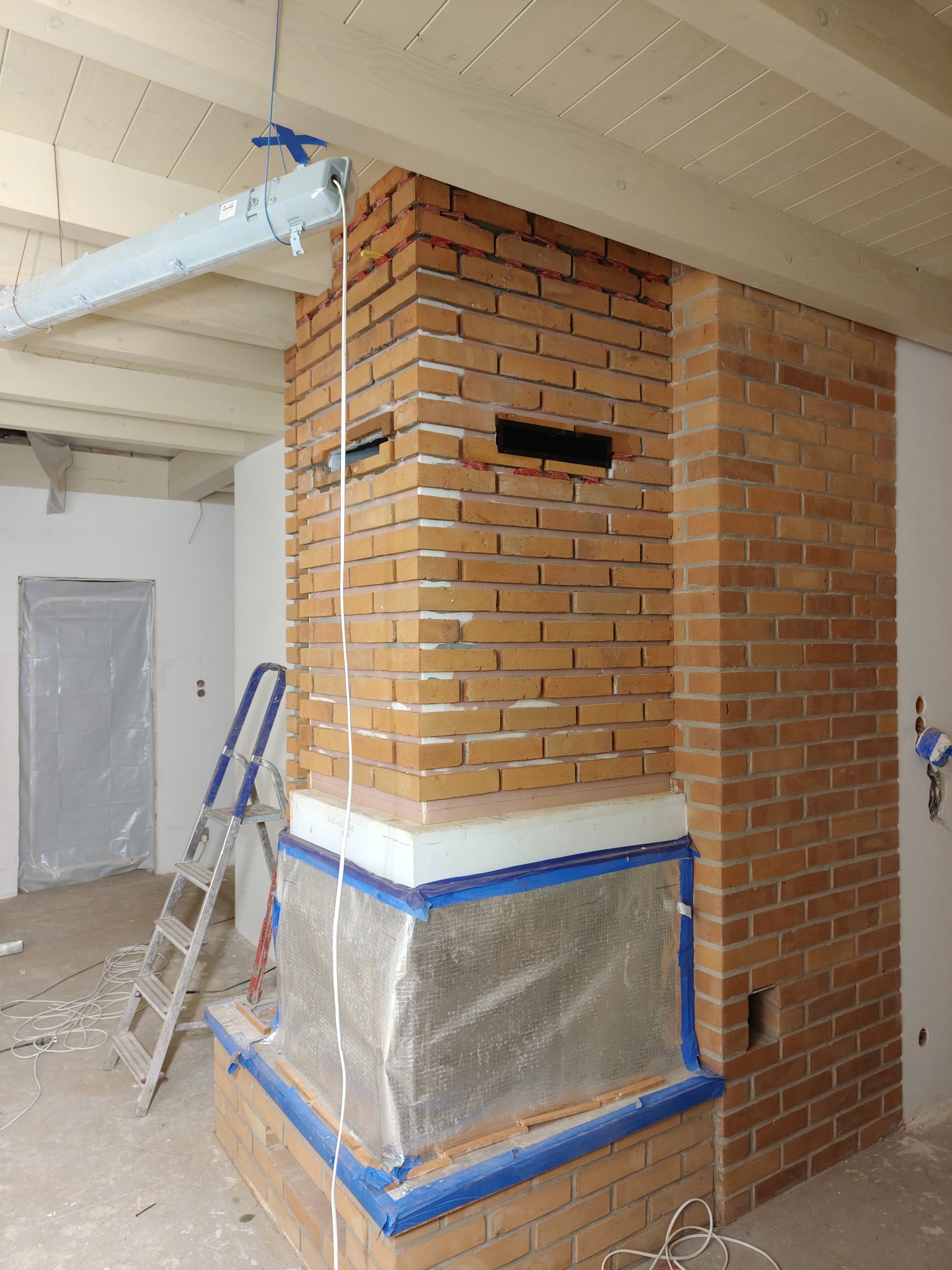 Murowany kominek z cegły w trakcie budowy, zabezpieczony folią i taśmą malarską, widoczna drabina i przewody elektryczne, surowy stan wykończenia wnętrza.