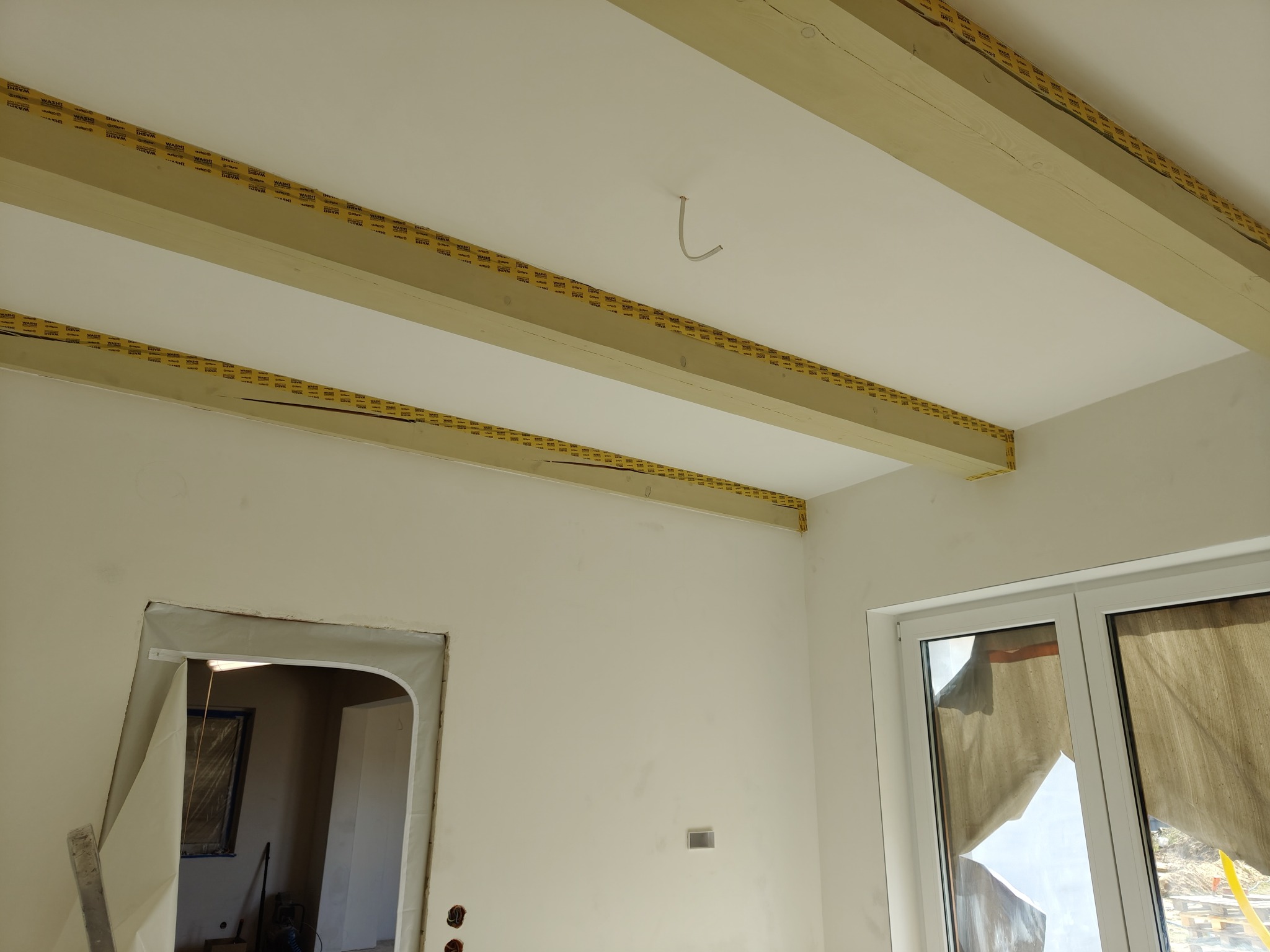 Wnętrze w trakcie prac wykończeniowych: widoczne drewniane belki stropowe zabezpieczone taśmą malarską, gładkie, białe ściany, zabezpieczone folią ościeżnice drzwiowe oraz okno z zasłoniętą szybą.