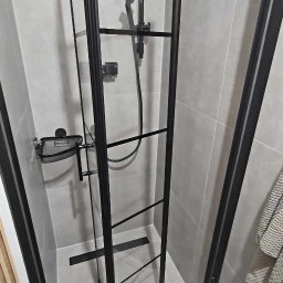 STJ - Usługi ogólnobudowlane Sylwia Fus - Nowoczesna kabina prysznicowa z czarnymi profilami i szklanymi drzwiami w wyremontowanej łazience, widoczny odpływ liniowy i płytki w odcieniach szarości.