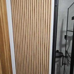 STJ - Usługi ogólnobudowlane Sylwia Fus - Ściana wykończona panelami z naturalnego drewna w pionowym układzie, widoczna kabina prysznicowa z czarnymi profilami w tle.