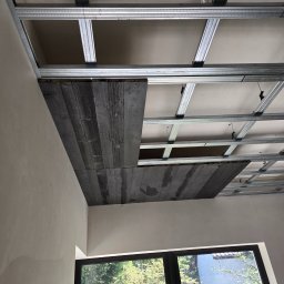 STJ - Usługi ogólnobudowlane Sylwia Fus - Montaż sufitu podwieszanego z płyt gipsowo-kartonowych na metalowej konstrukcji, częściowo wykończony deskami imitującymi stare drewno, widok z dołu na tle okna.