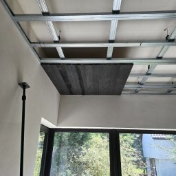 STJ - Usługi ogólnobudowlane Sylwia Fus - Konstrukcja metalowa sufitu podwieszanego z widocznym fragmentem drewnianej płyty, częściowo zakrywającej przestrzeń, w jasnym wnętrzu z oknem.