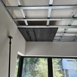 Konstrukcja metalowa sufitu podwieszanego z widocznym fragmentem drewnianej płyty, częściowo zakrywającej przestrzeń, w jasnym wnętrzu z oknem.