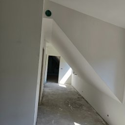 STJ - Usługi ogólnobudowlane Sylwia Fus - Poddasze w trakcie wykończenia: surowe ściany, przygotowane do malowania, widoczny otwór wentylacyjny i fragment okna dachowego z wpadającym światłem.