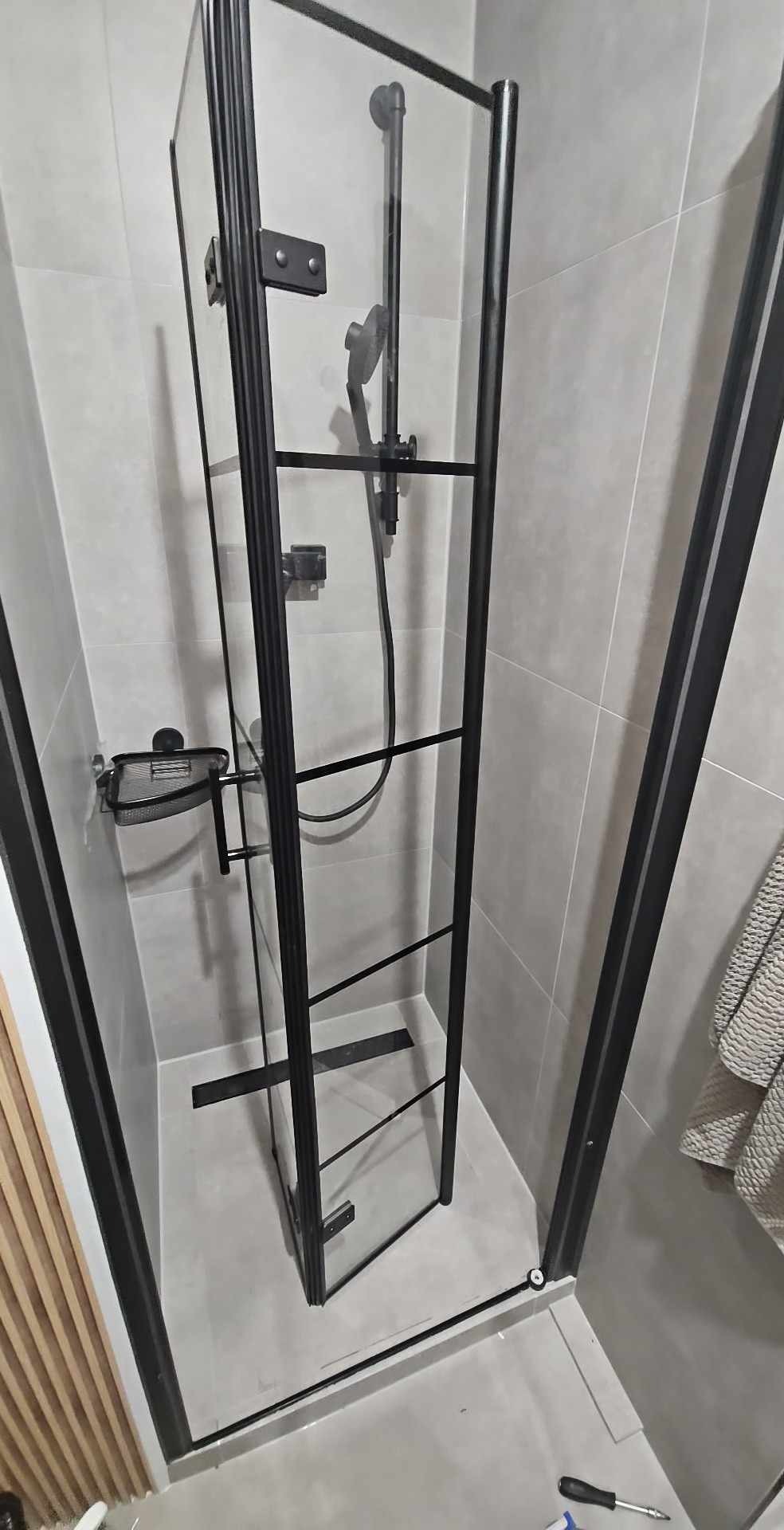 Nowoczesna kabina prysznicowa z czarnymi profilami i szklanymi drzwiami w wyremontowanej łazience, widoczny odpływ liniowy i płytki w odcieniach szarości.