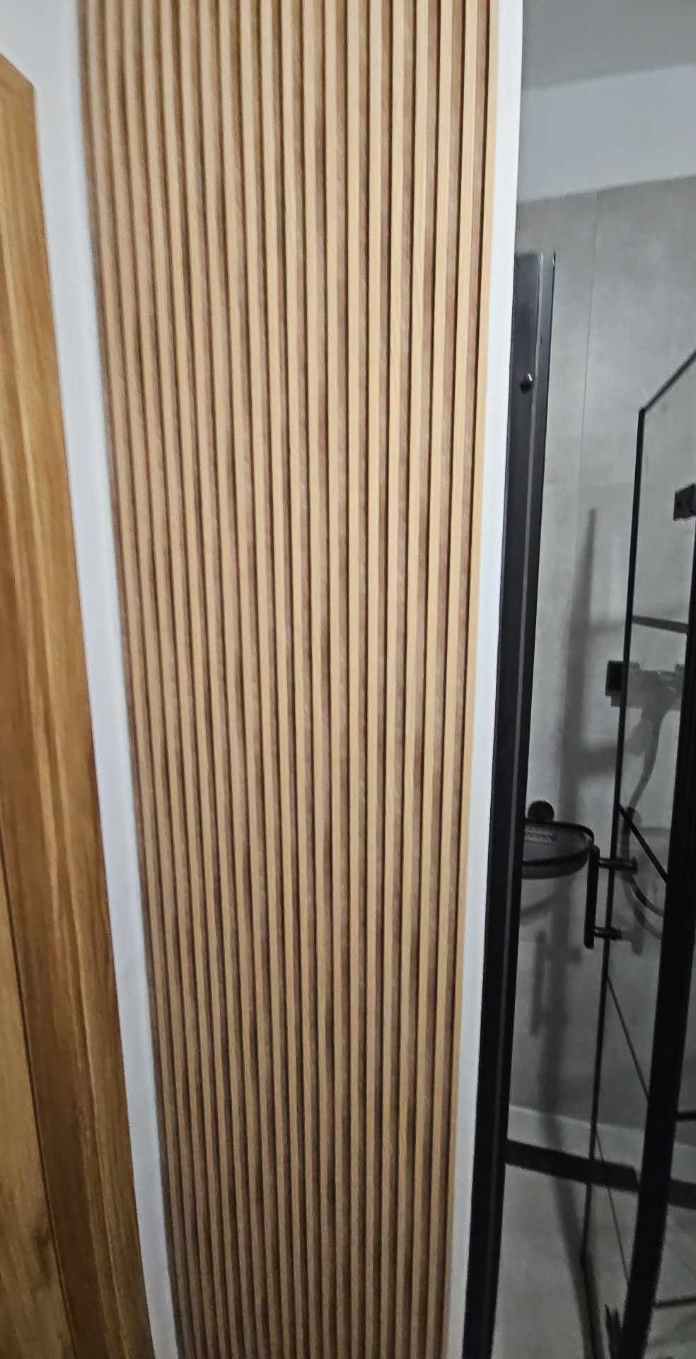 Ściana wykończona panelami z naturalnego drewna w pionowym układzie, widoczna kabina prysznicowa z czarnymi profilami w tle.