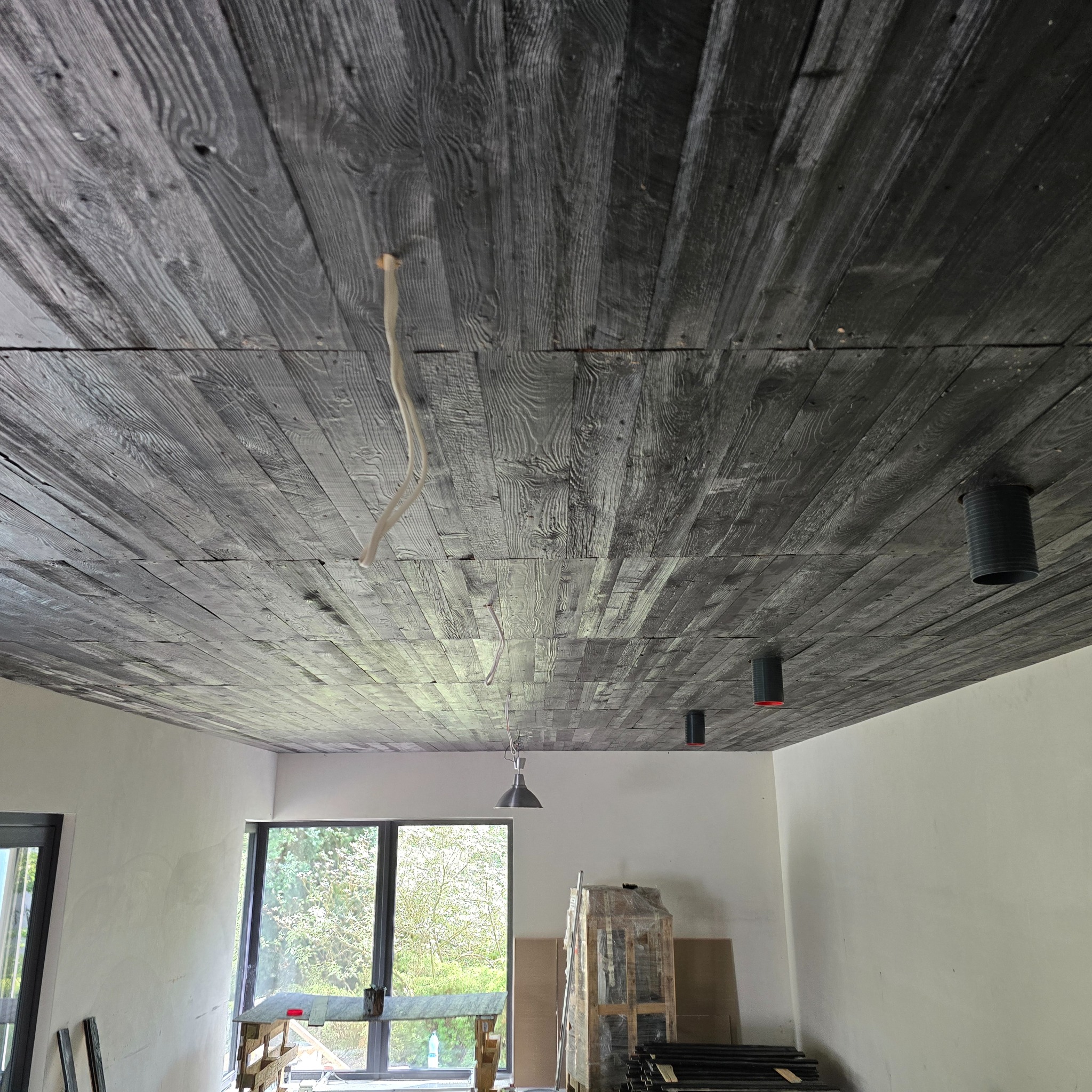 Drewniany sufit z widocznymi instalacjami elektrycznymi i oświetleniem, w trakcie wykańczania wnętrza, widoczne okna i elementy wyposażenia.