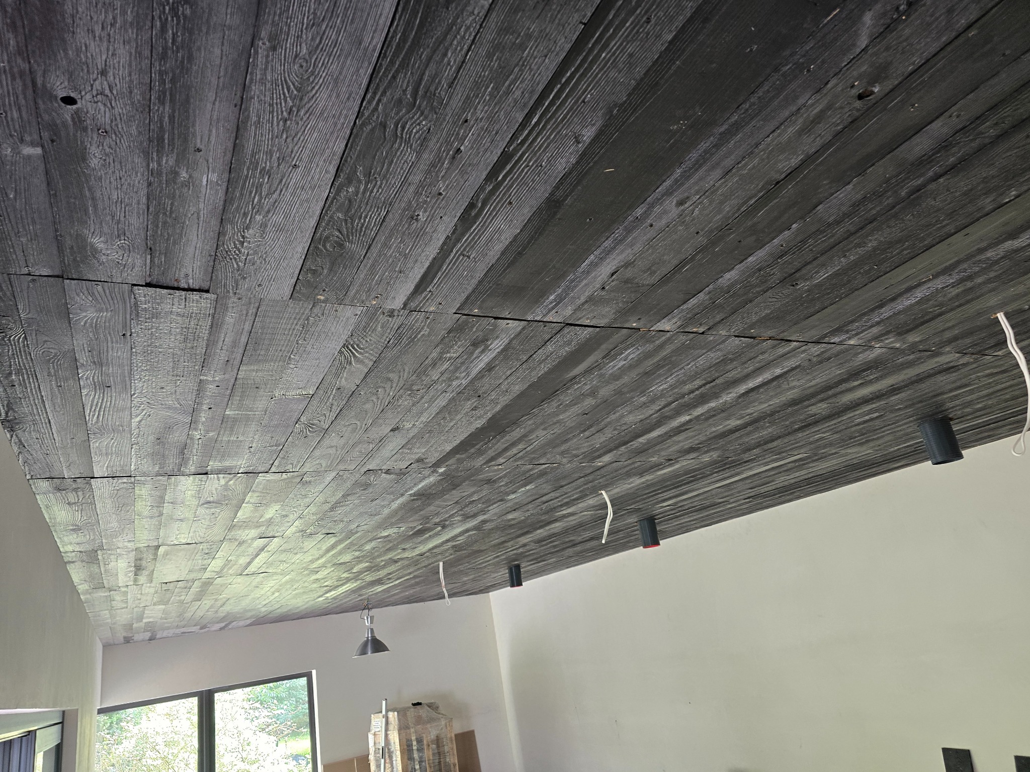 Drewniany sufit z widocznymi przewodami elektrycznymi i oprawami oświetleniowymi, w tle okno i część pomieszczenia w trakcie wykończenia.