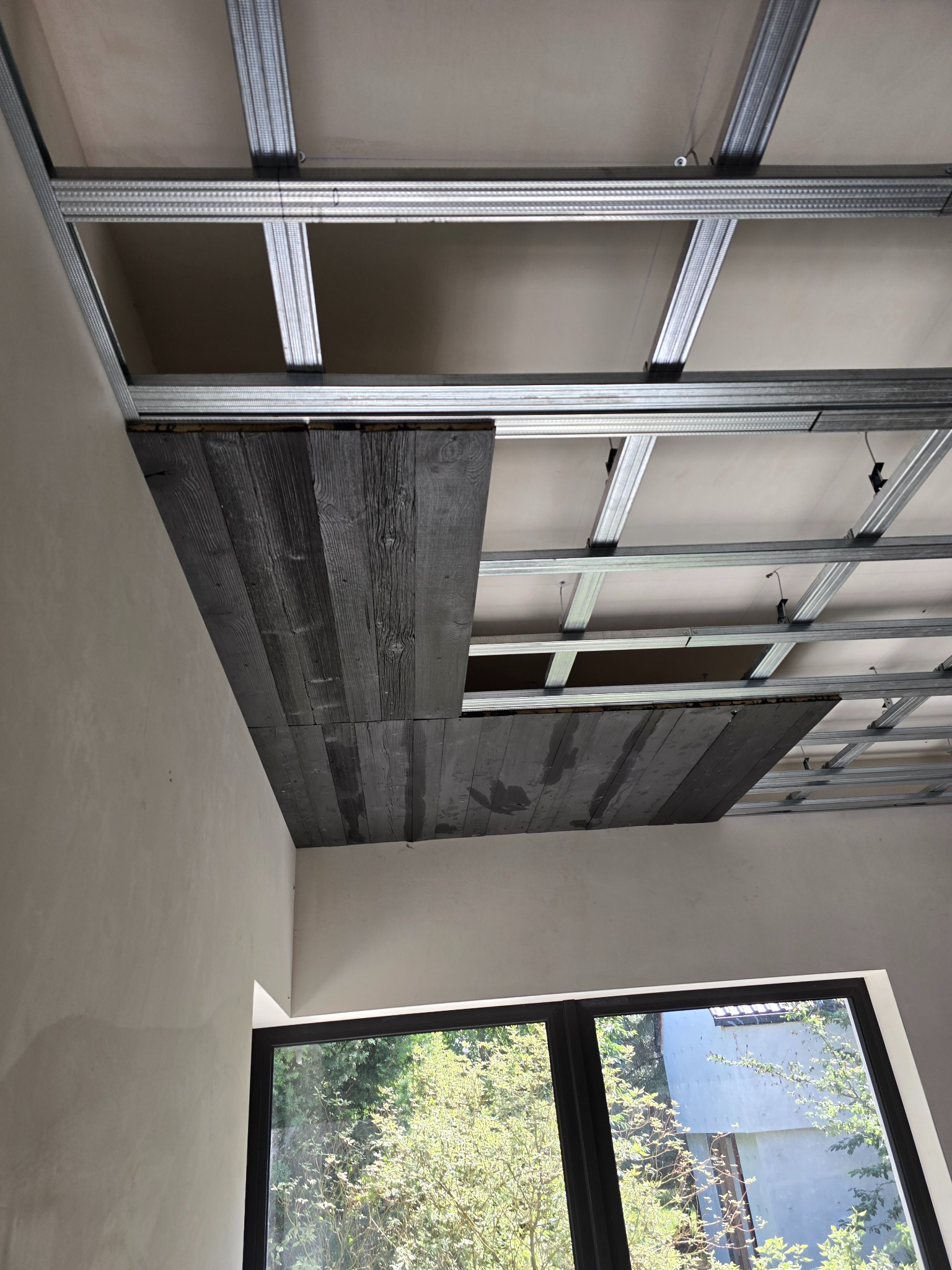 Montaż sufitu podwieszanego z płyt gipsowo-kartonowych na metalowej konstrukcji, częściowo wykończony deskami imitującymi stare drewno, widok z dołu na tle okna.