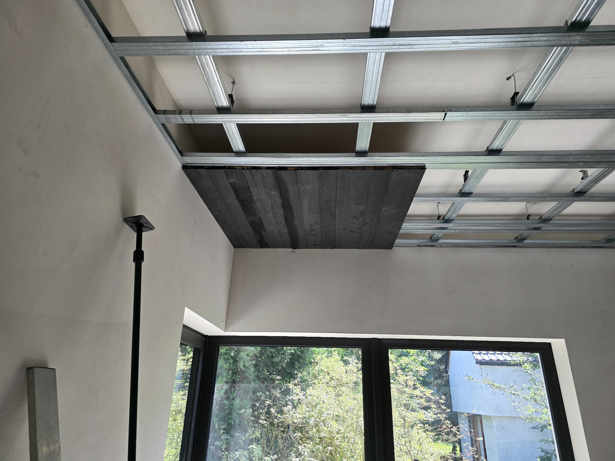 Konstrukcja metalowa sufitu podwieszanego z widocznym fragmentem drewnianej płyty, częściowo zakrywającej przestrzeń, w jasnym wnętrzu z oknem.