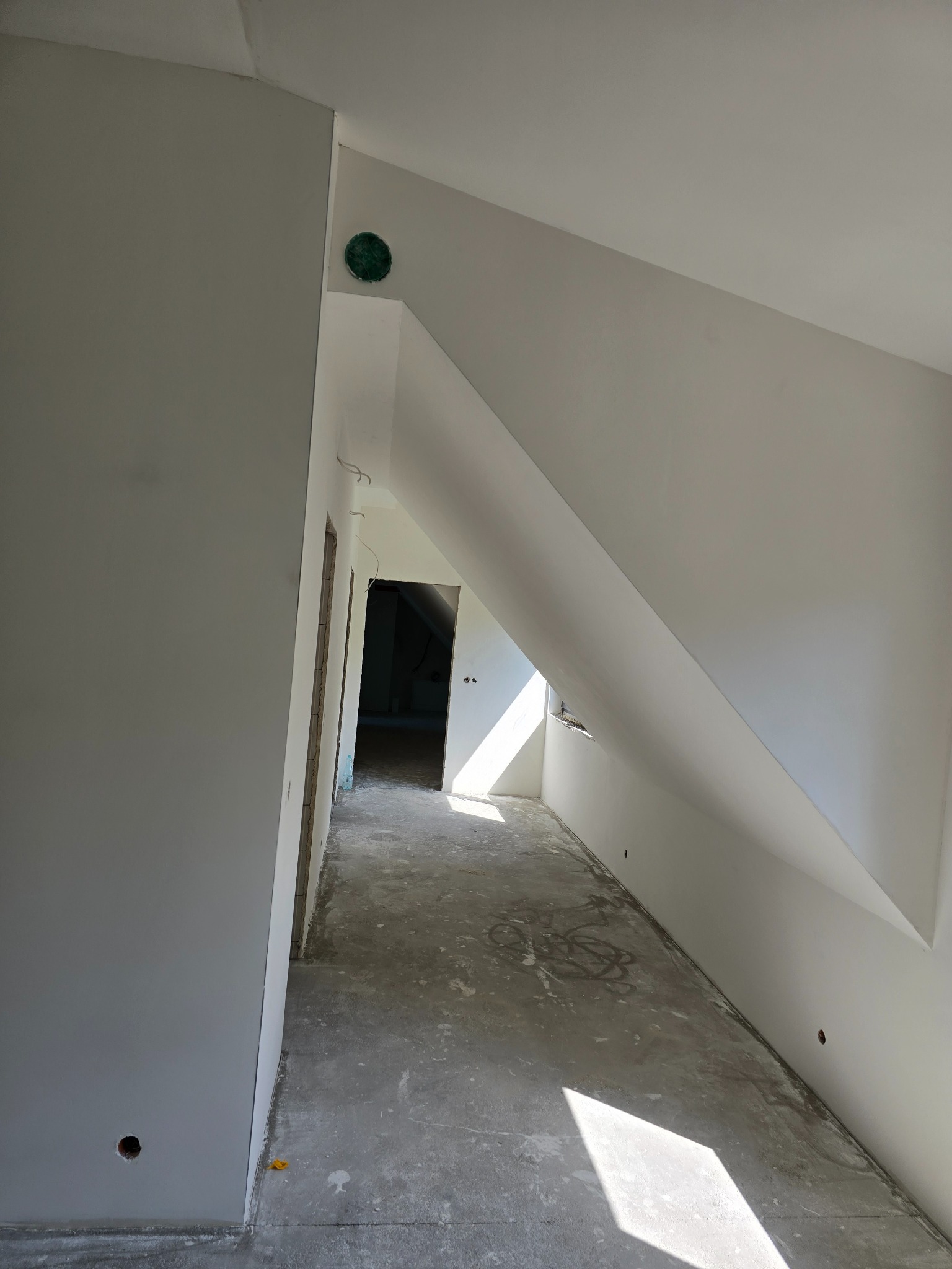 Poddasze w trakcie wykończenia: surowe ściany, przygotowane do malowania, widoczny otwór wentylacyjny i fragment okna dachowego z wpadającym światłem.