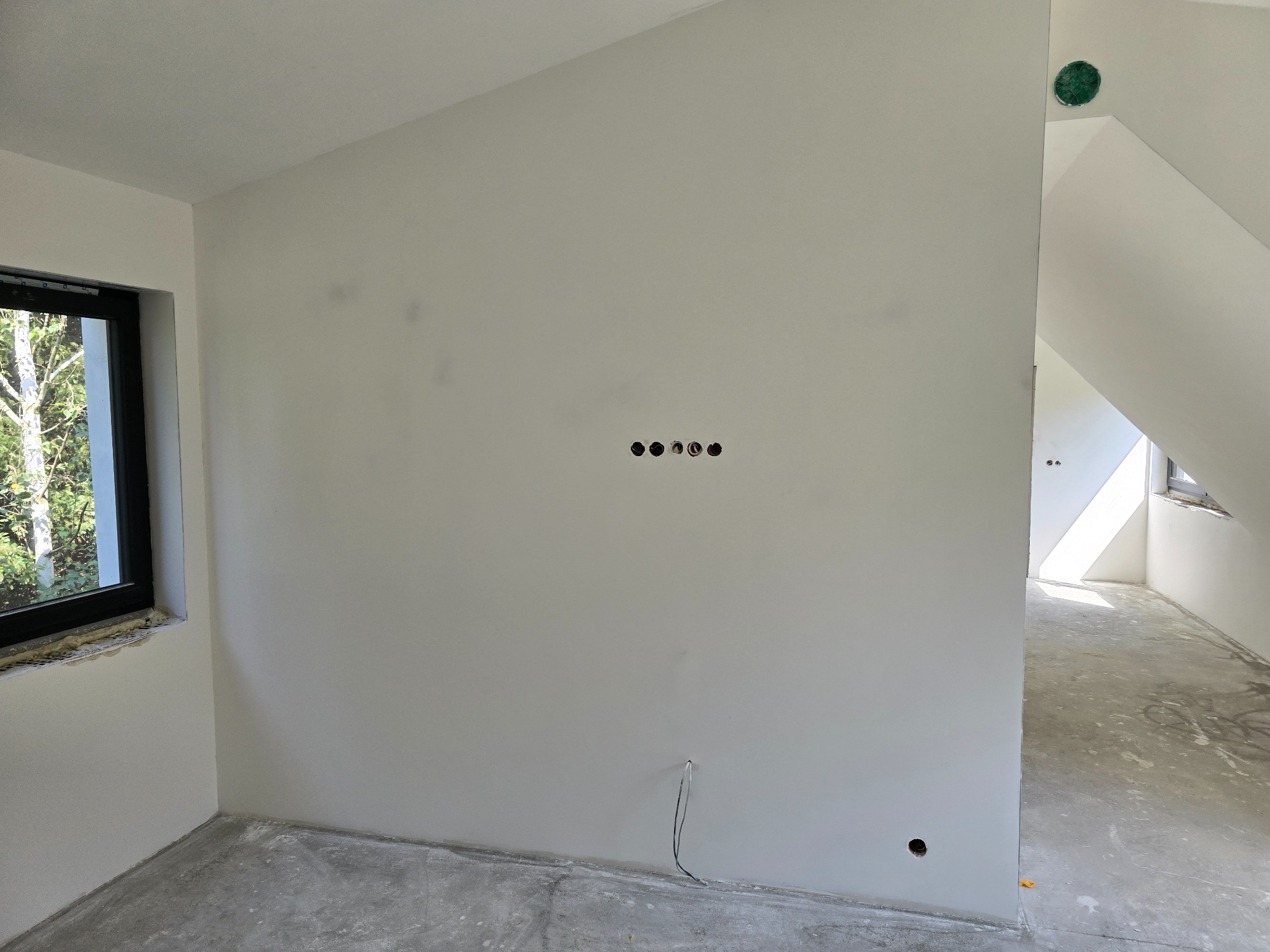 Poddasze w trakcie prac wykończeniowych: ściany przygotowane pod malowanie, widoczne otwory pod gniazdka elektryczne i wystający przewód, surowa wylewka na podłodze, okno w czarnej ramie.