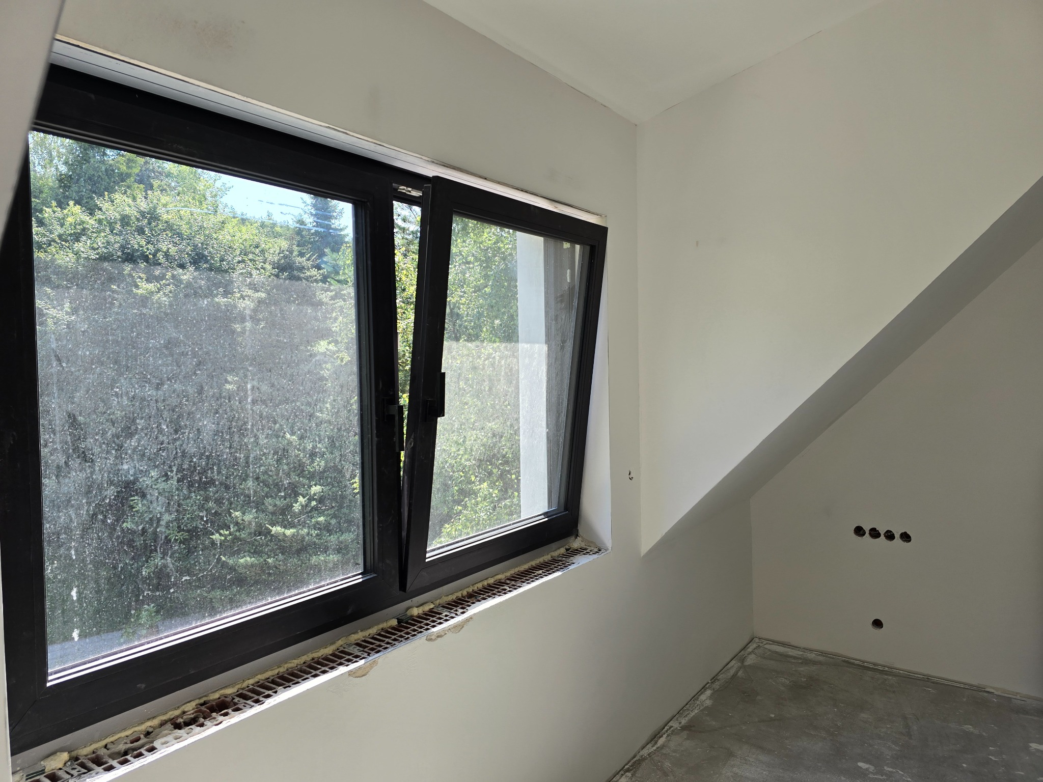 Poddasze w trakcie remontu: okno z czarną ramą, częściowo otwarte, widok na zieleń. Ściany gładkie, przygotowane do malowania. Widoczne puszki elektryczne.