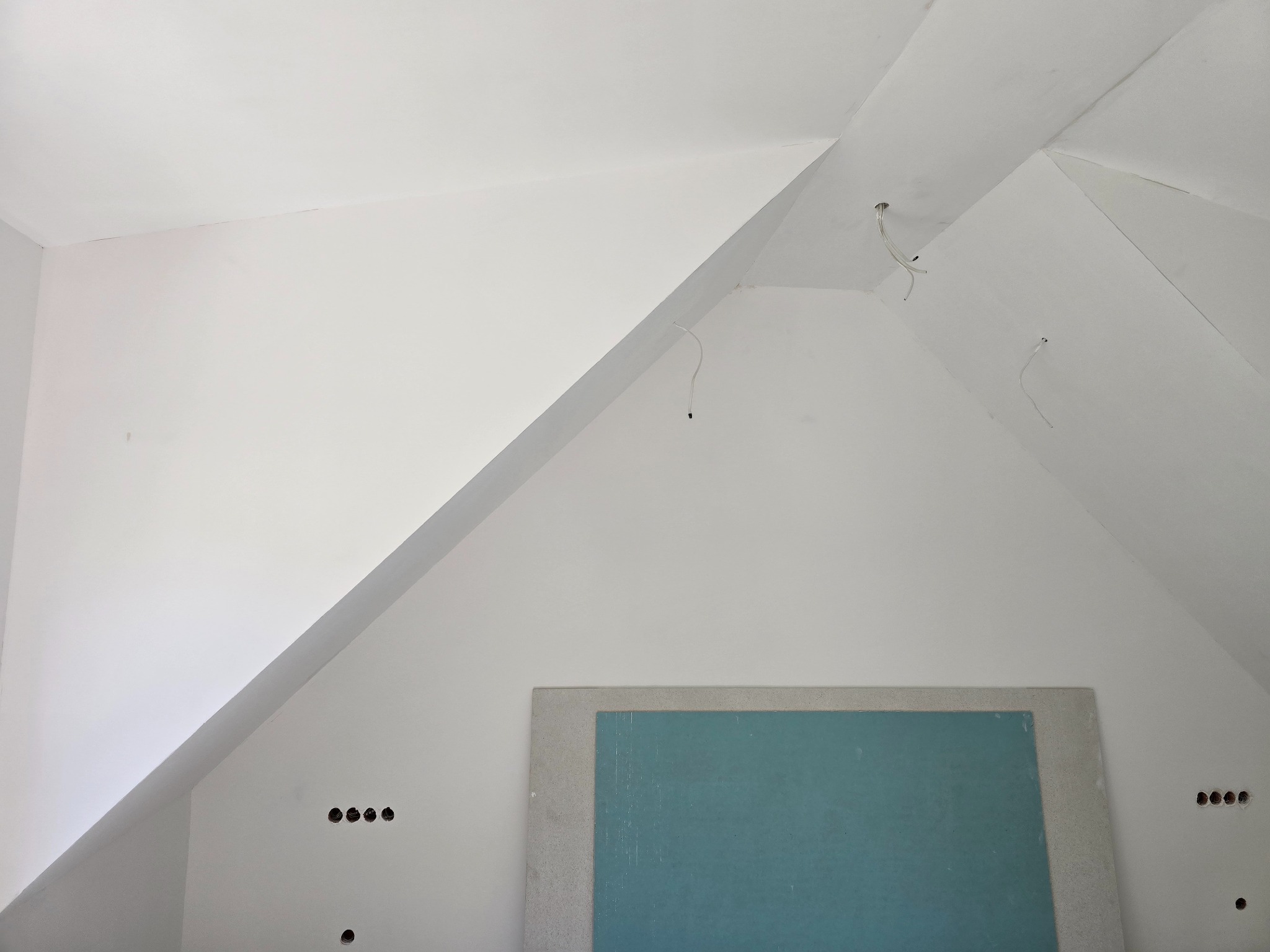 Poddasze po remoncie: białe ściany ze skosami, przygotowane otwory pod instalacje elektryczne, fragment zielonej płyty kartonowo-gipsowej.