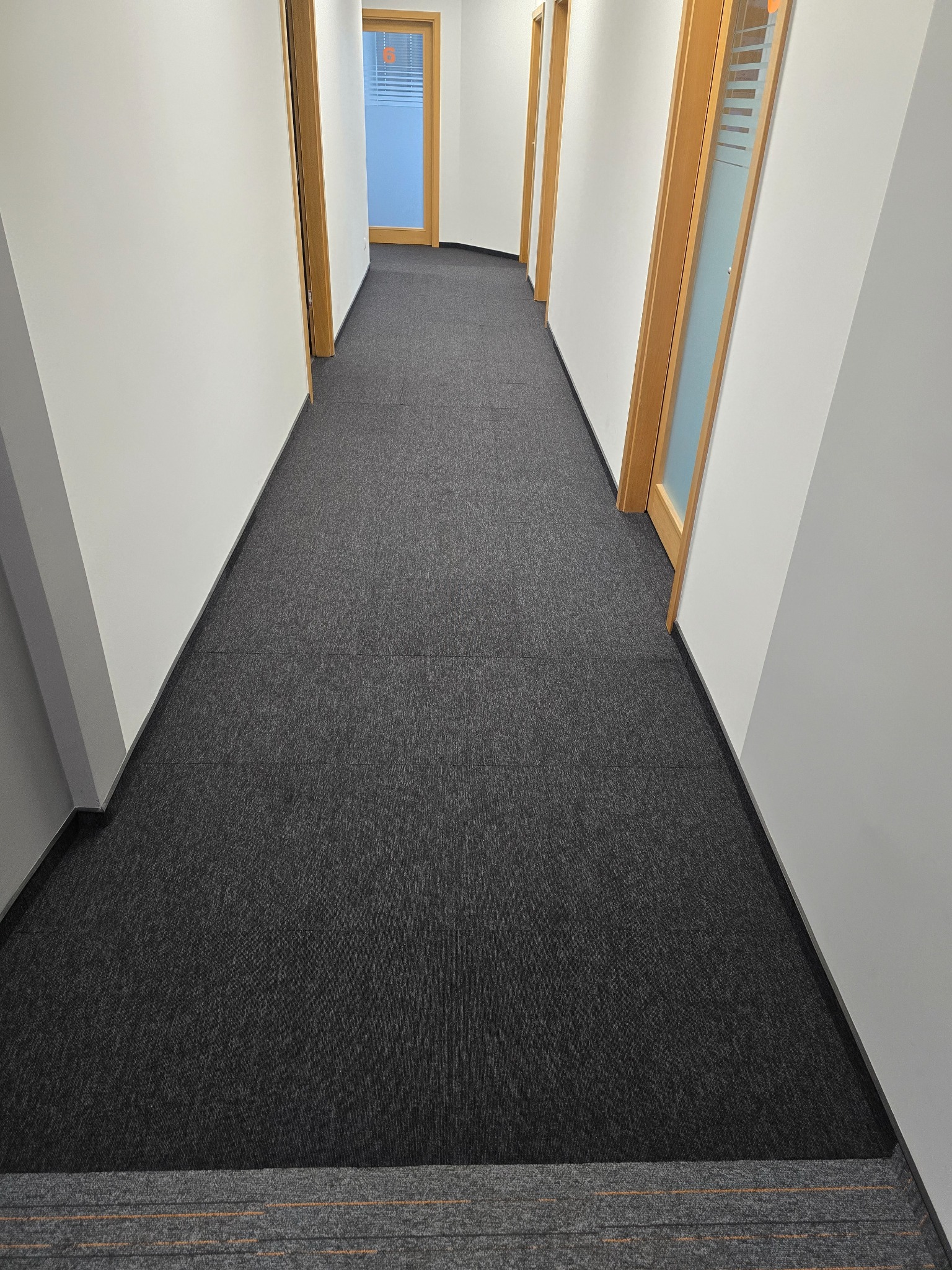 Długi korytarz biurowy z szarą wykładziną dywanową w płytkach, białymi ścianami i drzwiami z drewnianymi ościeżnicami i częściowo przeszklonymi szybami.