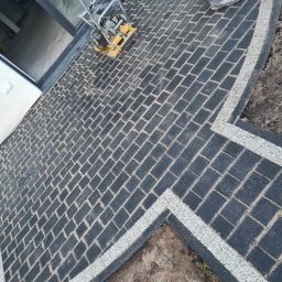Leobud - Ułożona czarna kostka brukowa z jasnymi fugami i obramowaniem, zagęszczarka wibracyjna obok drzwi, widoczne buty pracownika.