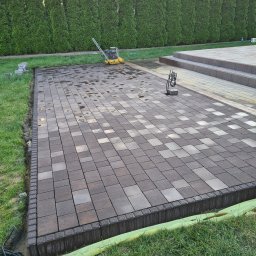 Leobud - Ułożona, mokra kostka brukowa w odcieniach szarości i brązu z widocznym obrzeżem, ubijarka wibracyjna oraz zielony trawnik i tuje w tle.