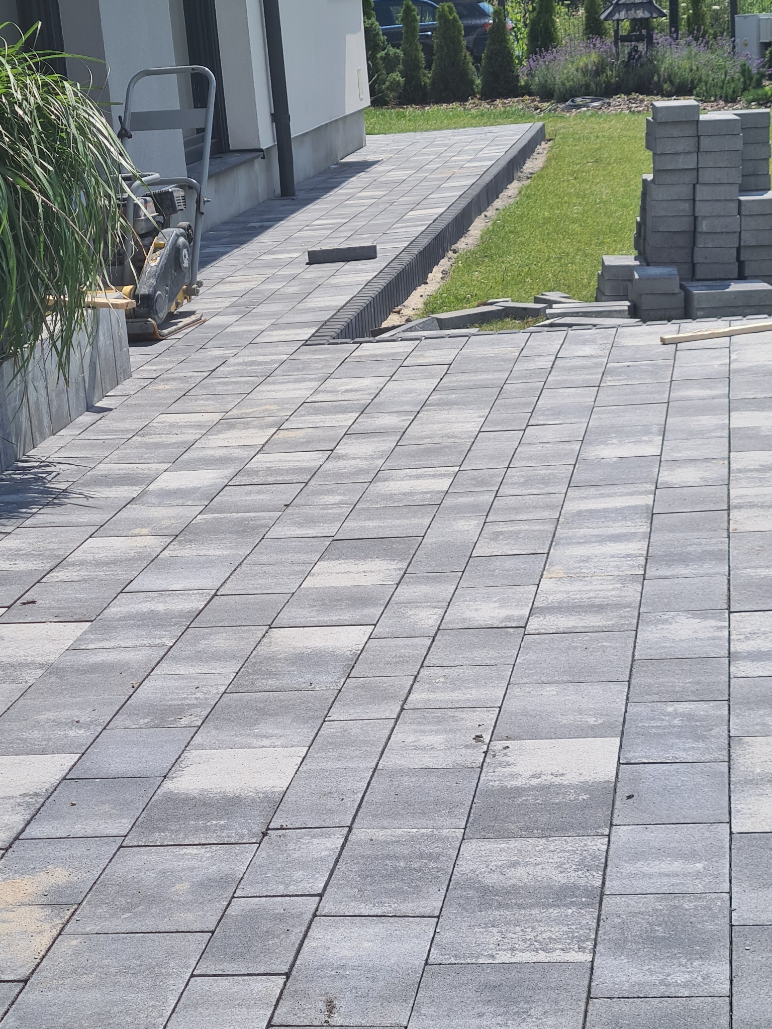 Ułożona szara kostka brukowa na posesji z widocznym ubijakiem wibracyjnym, stosem kostki i trawnikiem w tle.