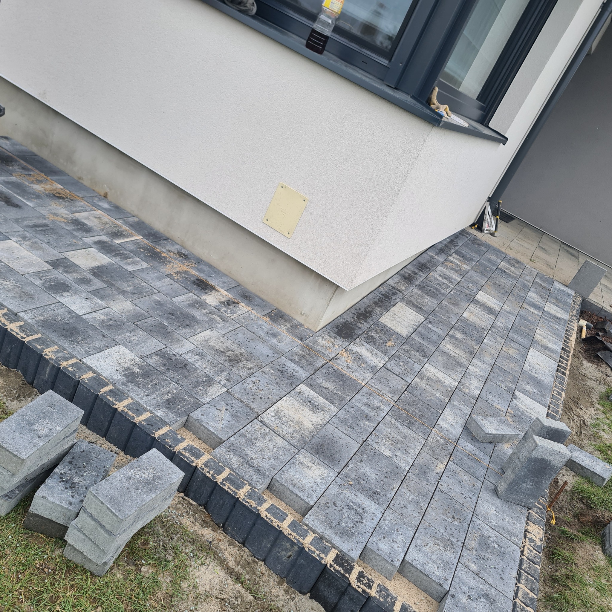 Fragment posesji z częściowo ułożoną szarą kostką brukową i czarnym obrzeżem, widoczne zapasy kostki oraz fragment budynku z oknem.