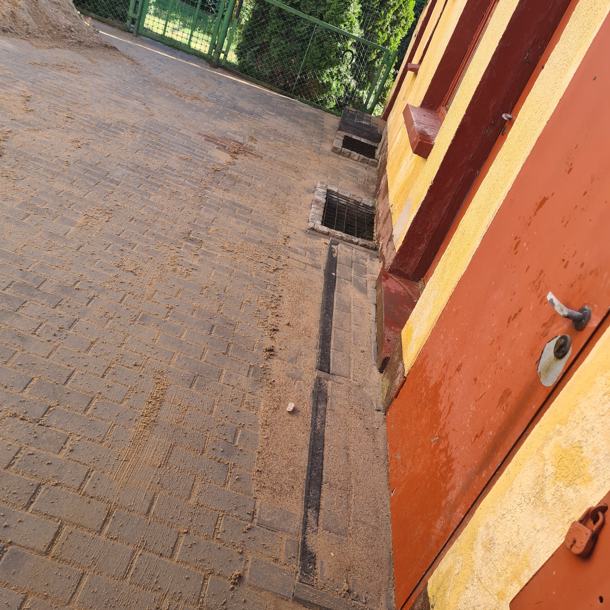 Ułożona kostka brukowa pokryta piaskiem, widoczne studzienki ściekowe i fragment pomarańczowej ściany budynku z metalowymi drzwiami.