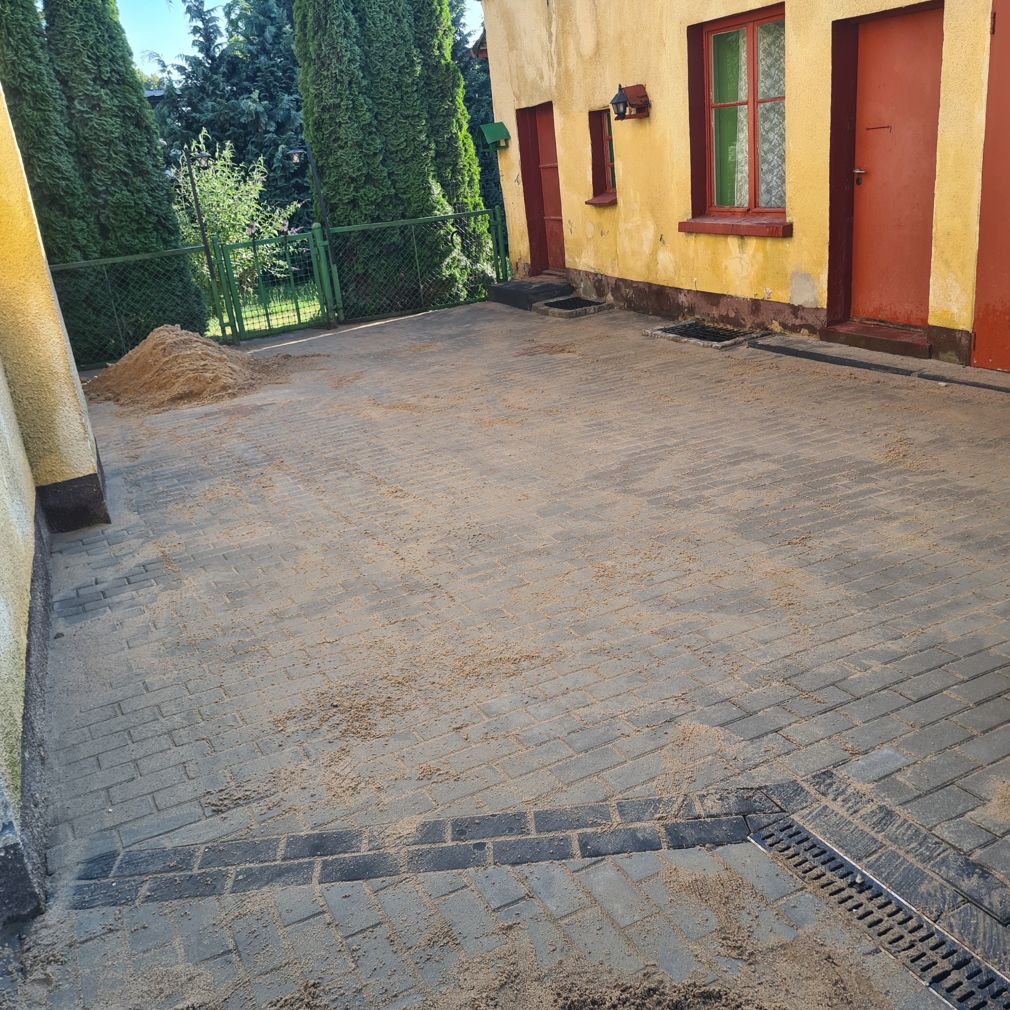 Szara kostka brukowa ułożona na podwórku, zasypana piaskiem, z widocznym odpływem liniowym i stertą piasku przy budynku z czerwonymi drzwiami i oknami.