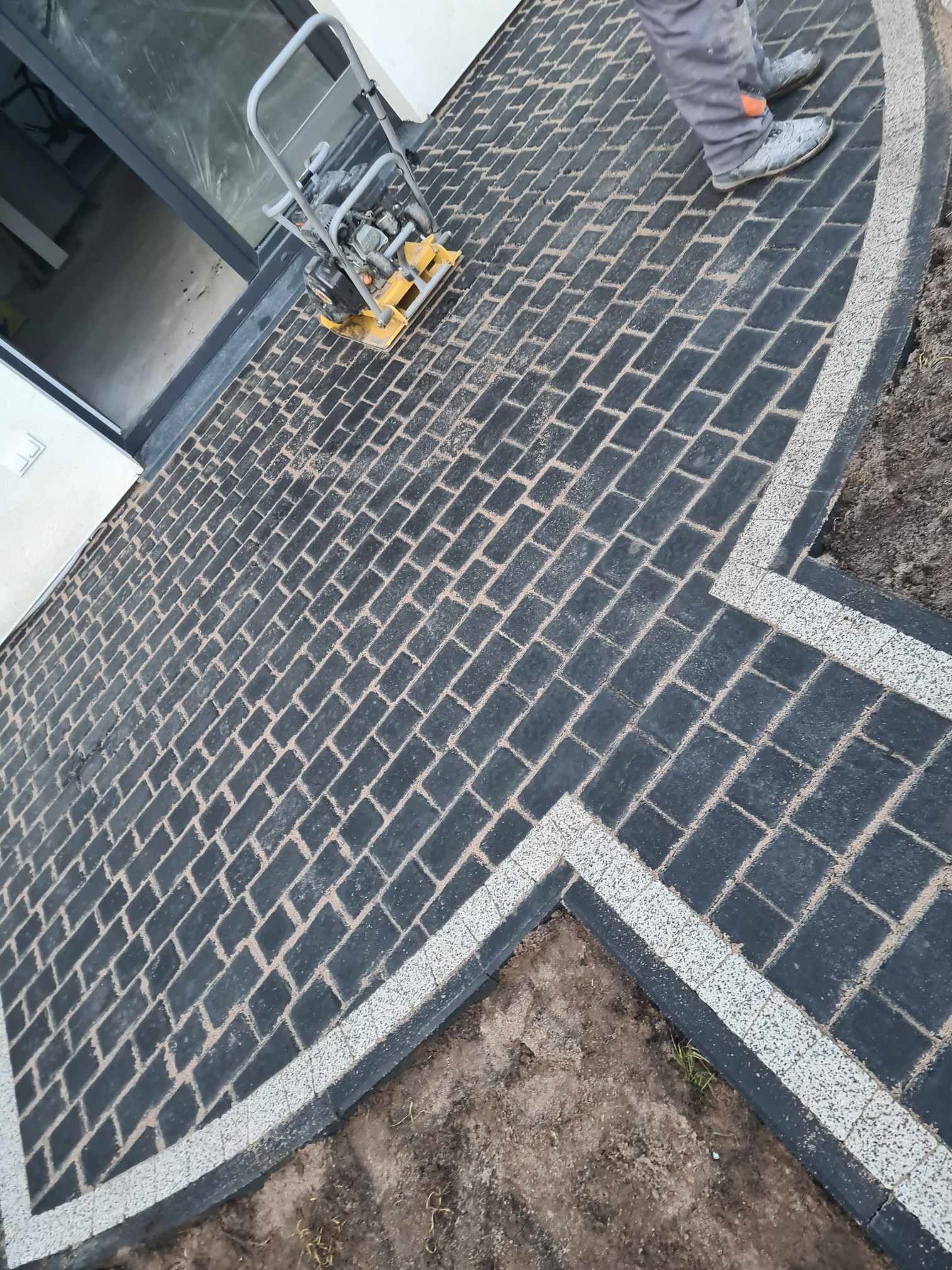 Ułożona czarna kostka brukowa z jasnymi fugami i obramowaniem, zagęszczarka wibracyjna obok drzwi, widoczne buty pracownika.