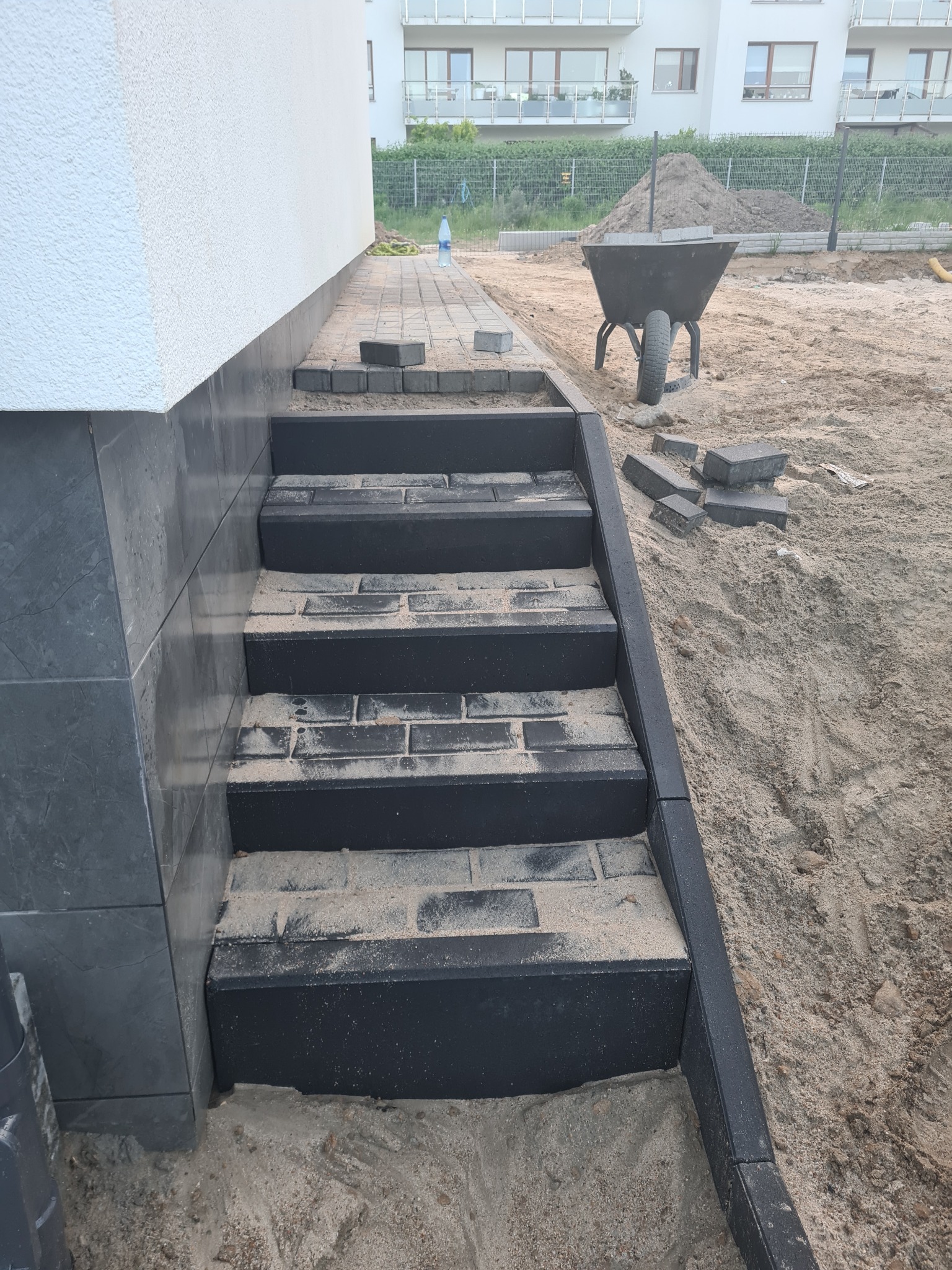Schody z ciemnej kostki brukowej w trakcie budowy, z widocznym piaskiem na stopniach i wokół, taczka z ziemią w tle.
