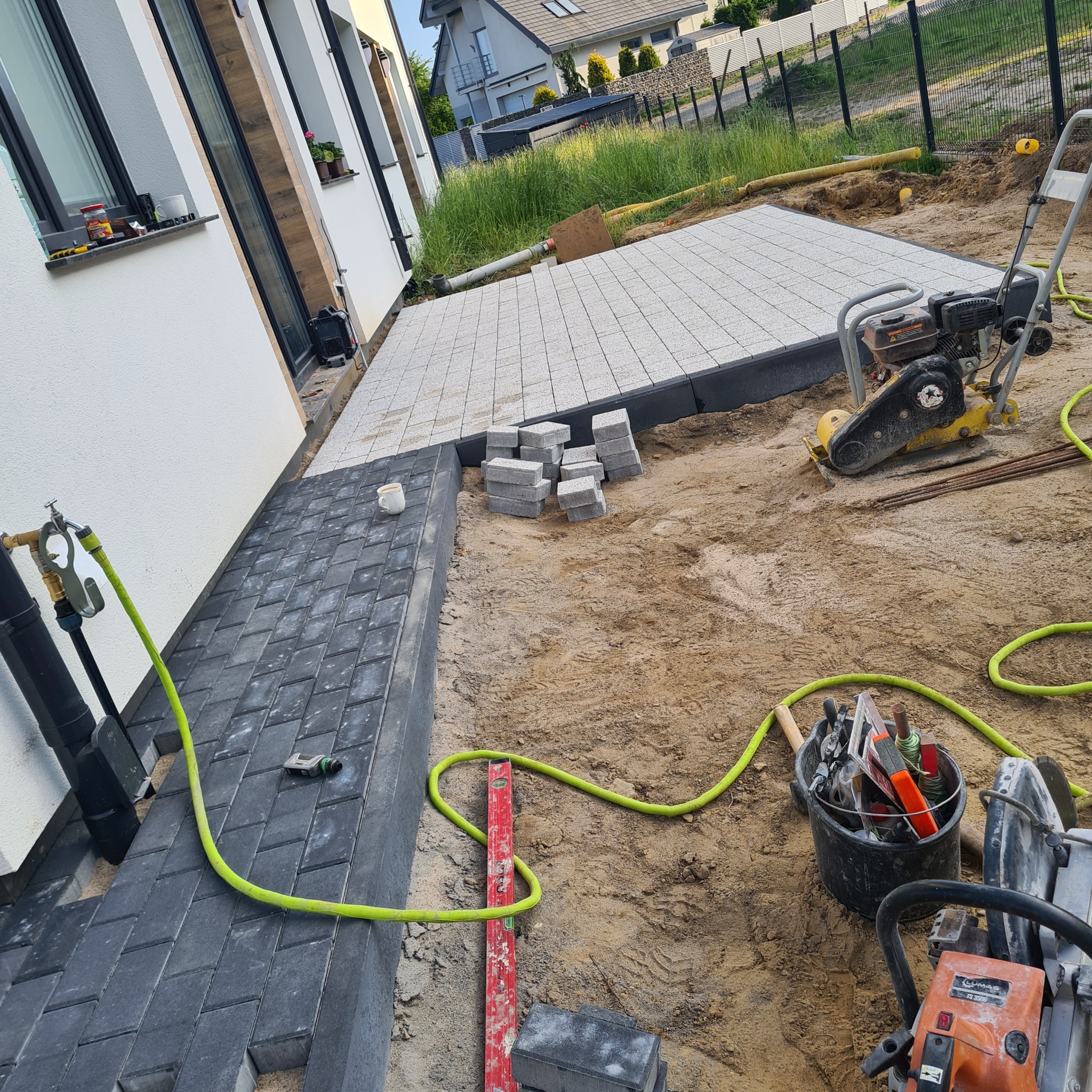 Fragment układanej ścieżki z ciemnej kostki obok białego budynku, z przygotowanym podłożem pod jasną kostkę, ubijarką, stosem kostki, poziomnicą i narzędziami w wiadrze.