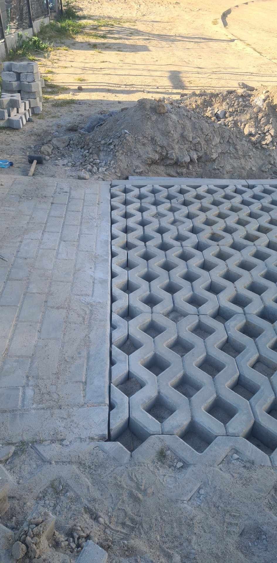 Fragment powierzchni z ułożoną szarą kostką ażurową o geometrycznym wzorze, sąsiadującą z prostokątną kostką, widoczny stos kostki i pryzma piasku w tle.