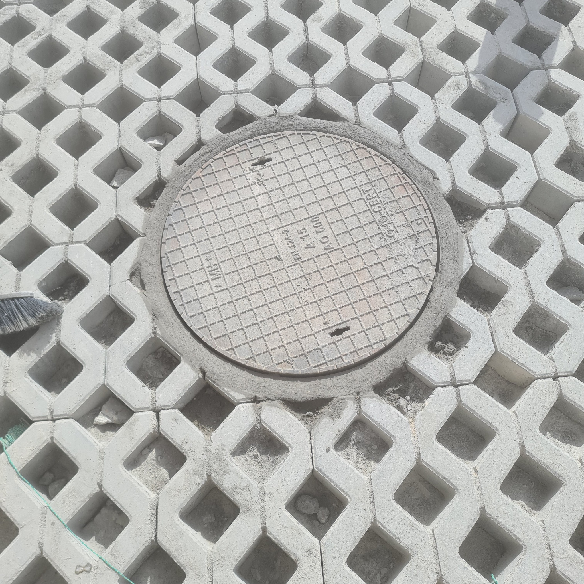 Szara ażurowa kostka betonowa typu plaster miodu z okrągłą studzienką kanalizacyjną pośrodku, wypełniona piaskiem, z widocznym fragmentem szczotki do zamiatania.