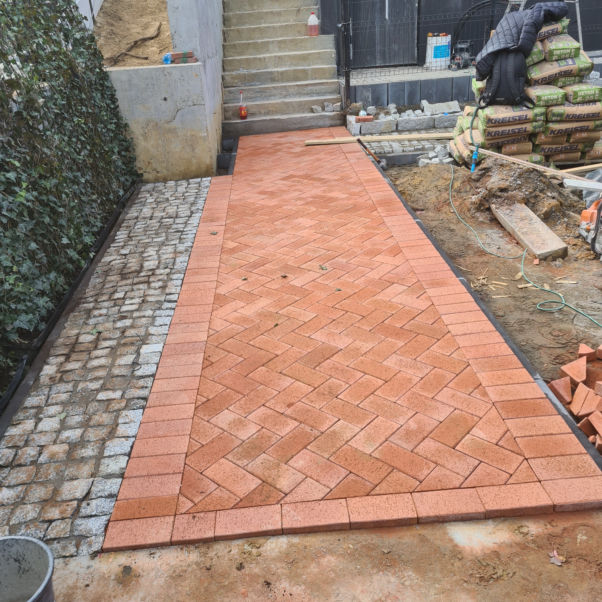 Ścieżka z czerwonej kostki ułożonej w jodełkę z obramowaniem, częściowo obok istniejącej nawierzchni z szarego kamienia, widok z góry, obok schody i worki z betonem.