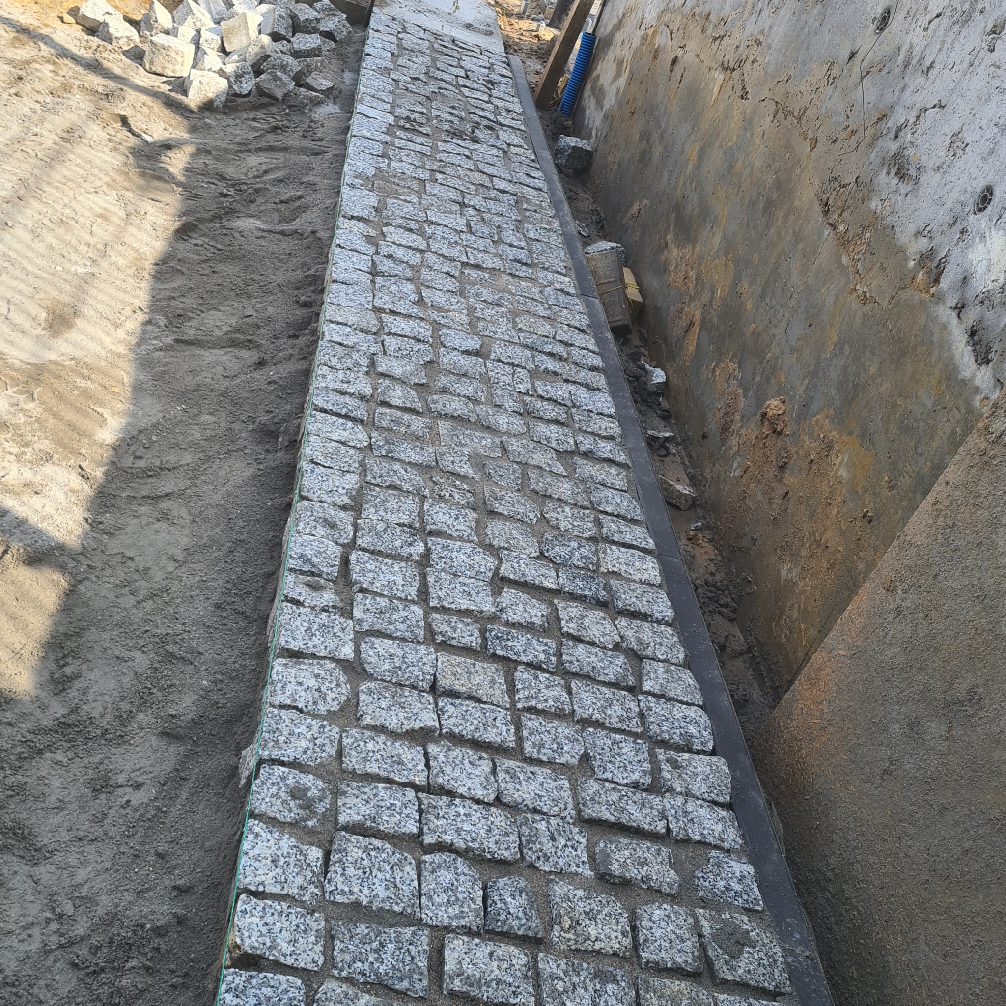 Fragment nowo ułożonej ścieżki z kostki granitowej o nieregularnych kształtach, widoczny piasek po bokach i betonowa ściana wykopu, z zaznaczonymi liniami poziomymi.