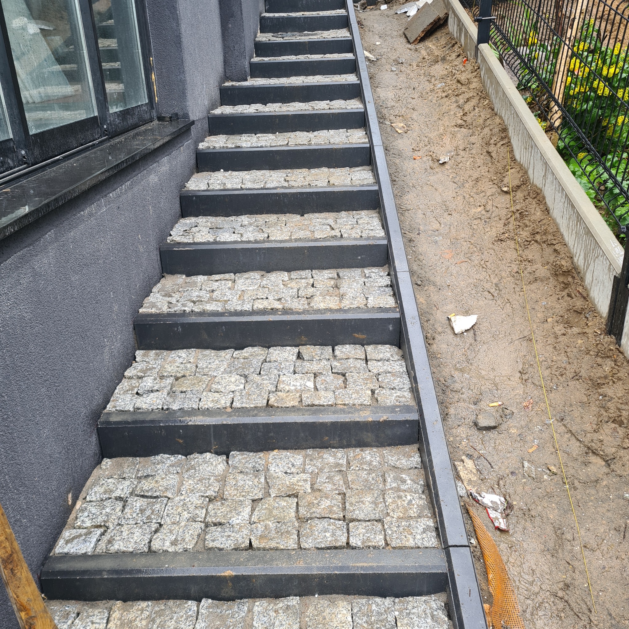 Schody zewnętrzne z kostki brukowej w odcieniach szarości, z czarnymi obrzeżami, prowadzące w górę, obok budynku z szarą elewacją i okna, oraz ogrodzenia z ziemistym podłożem.
