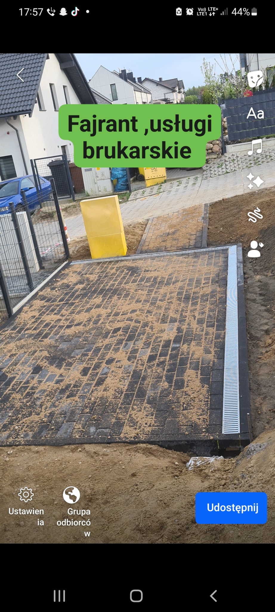 Ułożona kostka brukowa na posesji, zasypana piaskiem kwarcowym, widoczny system odprowadzania wody, fragment ogrodzenia i budynek w tle.