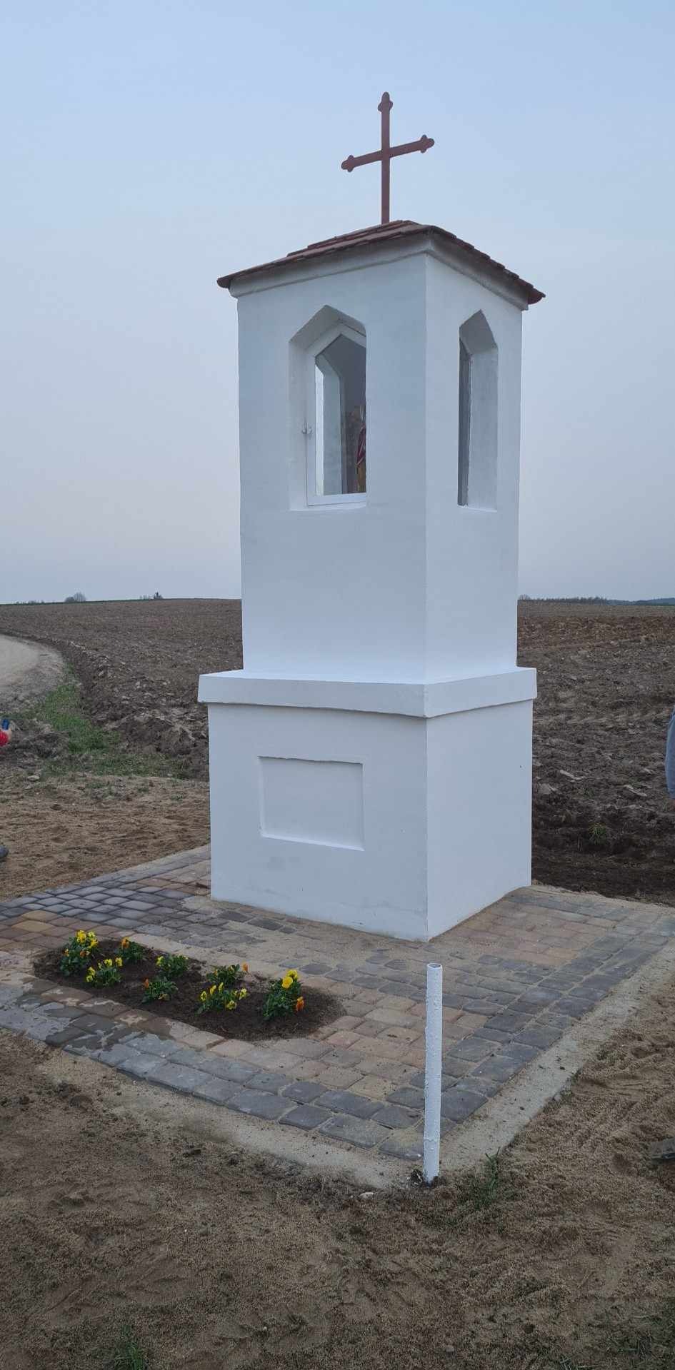Biała kapliczka z krzyżem na szczycie, otoczona świeżo ułożoną kostką brukową i rabatką z żółtymi kwiatami, na tle zaoranego pola.