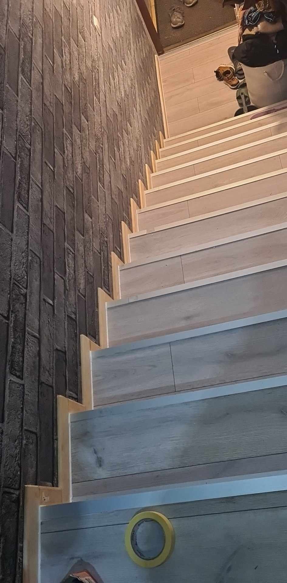 Wykończone schody z drewnopodobnymi panelami, zabezpieczone taśmą malarską, z dekoracyjną ścianą z cegły w tle.