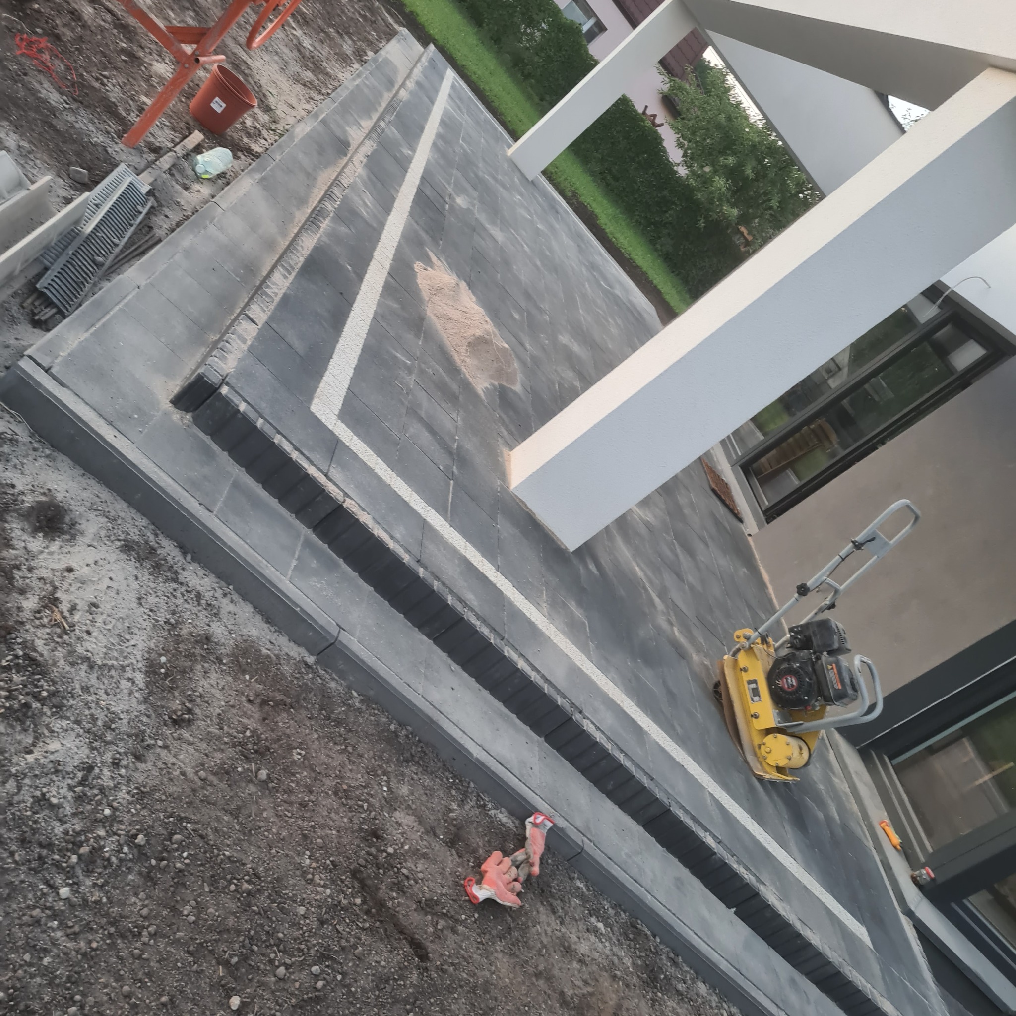 Układanie kostki brukowej w nowoczesnym stylu z białym pasem dekoracyjnym, ubijarka wibracyjna na powierzchni, obrzeża chodnikowe i robocze rękawice w tle.