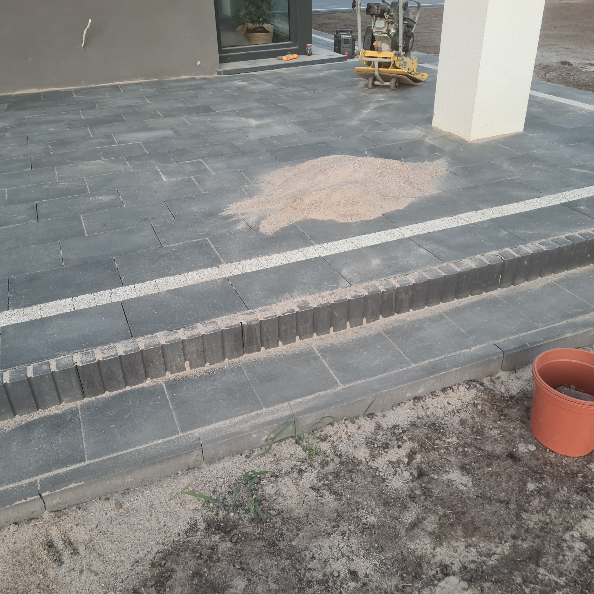 Ułożona, szara kostka brukowa z białymi fugami i obrzeżami, widoczny ubijak wibracyjny i rozsypany piasek kwarcowy, brązowa doniczka przy krawędzi.