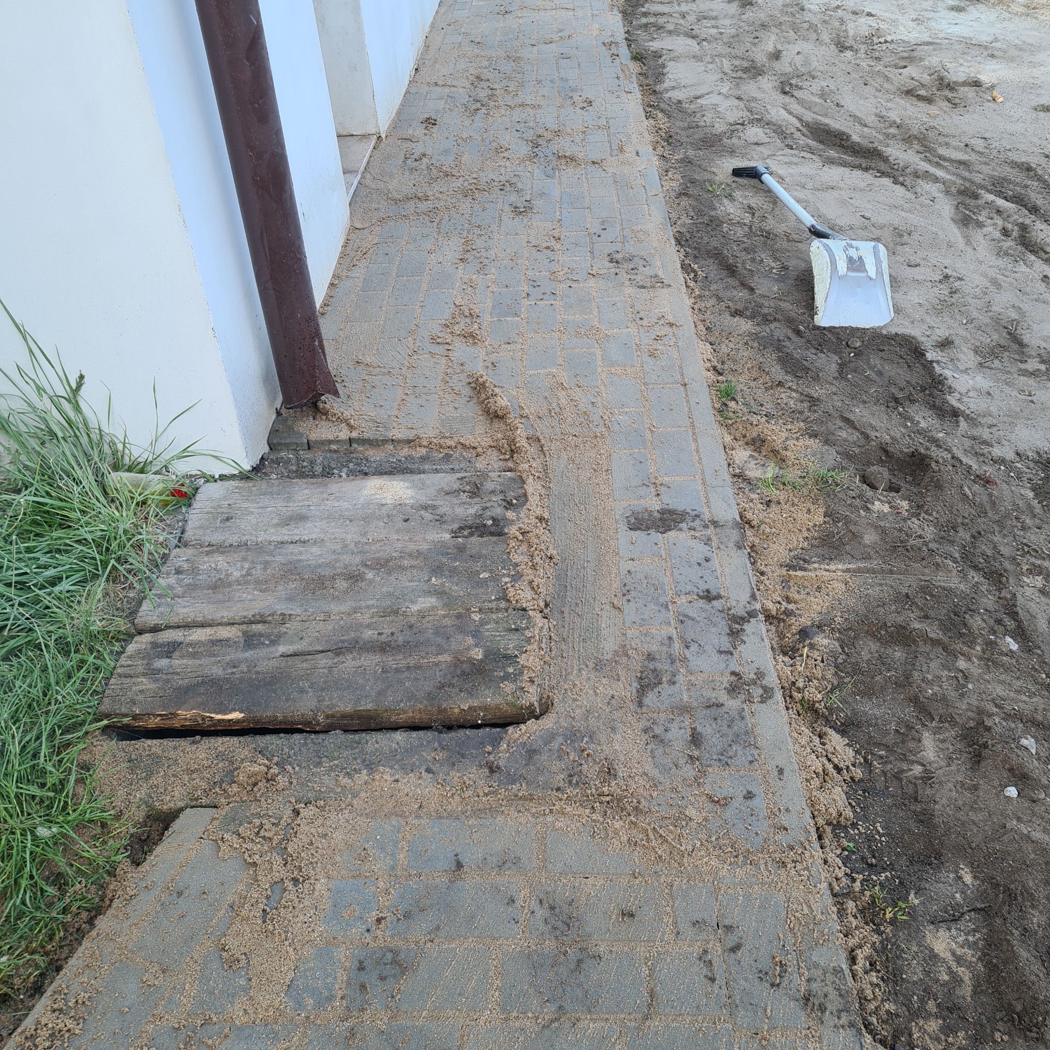 Szara kostka brukowa częściowo zasypana piaskiem, z widoczną drewnianą kładką przy ścianie budynku i łopatą na ziemi obok.