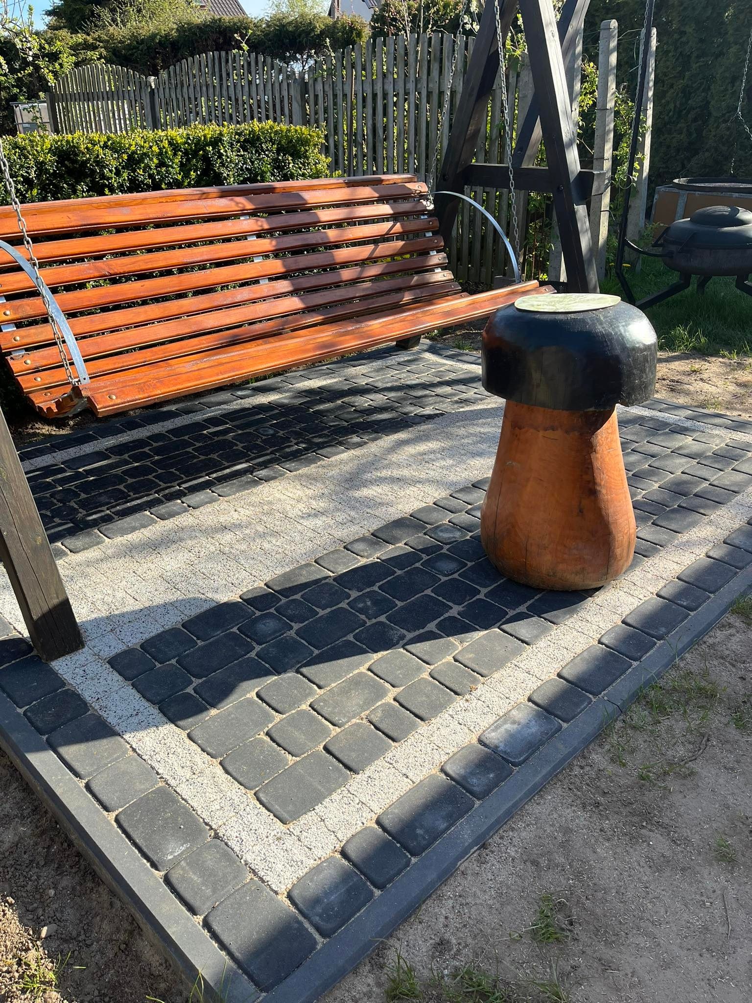 Drewniana huśtawka ogrodowa z siedziskiem na łańcuchach, umieszczona na nawierzchni z kostki brukowej w dwóch kolorach, obok drewniany stołek w kształcie grzyba.