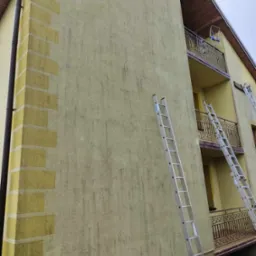 Żółta elewacja domu wielorodzinnego przed malowaniem, widoczne ślady zabrudzeń i dwie aluminiowe drabiny Krause oparte o ścianę.