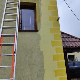 cleanup Karolina Ogrodnik - Fragment elewacji budynku z drabina opartą o ścianę, widoczne świeżo nałożone pasy farby w odcieniach żółci, okno z brązową ramą i fragment dachu sąsiedniego budynku.
