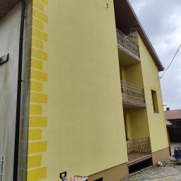 cleanup Karolina Ogrodnik - Żółta elewacja domu z dekoracyjnymi elementami w kształcie cegieł na rogu, widoczne balkony z metalowymi balustradami i narzędzia na kostce brukowej.