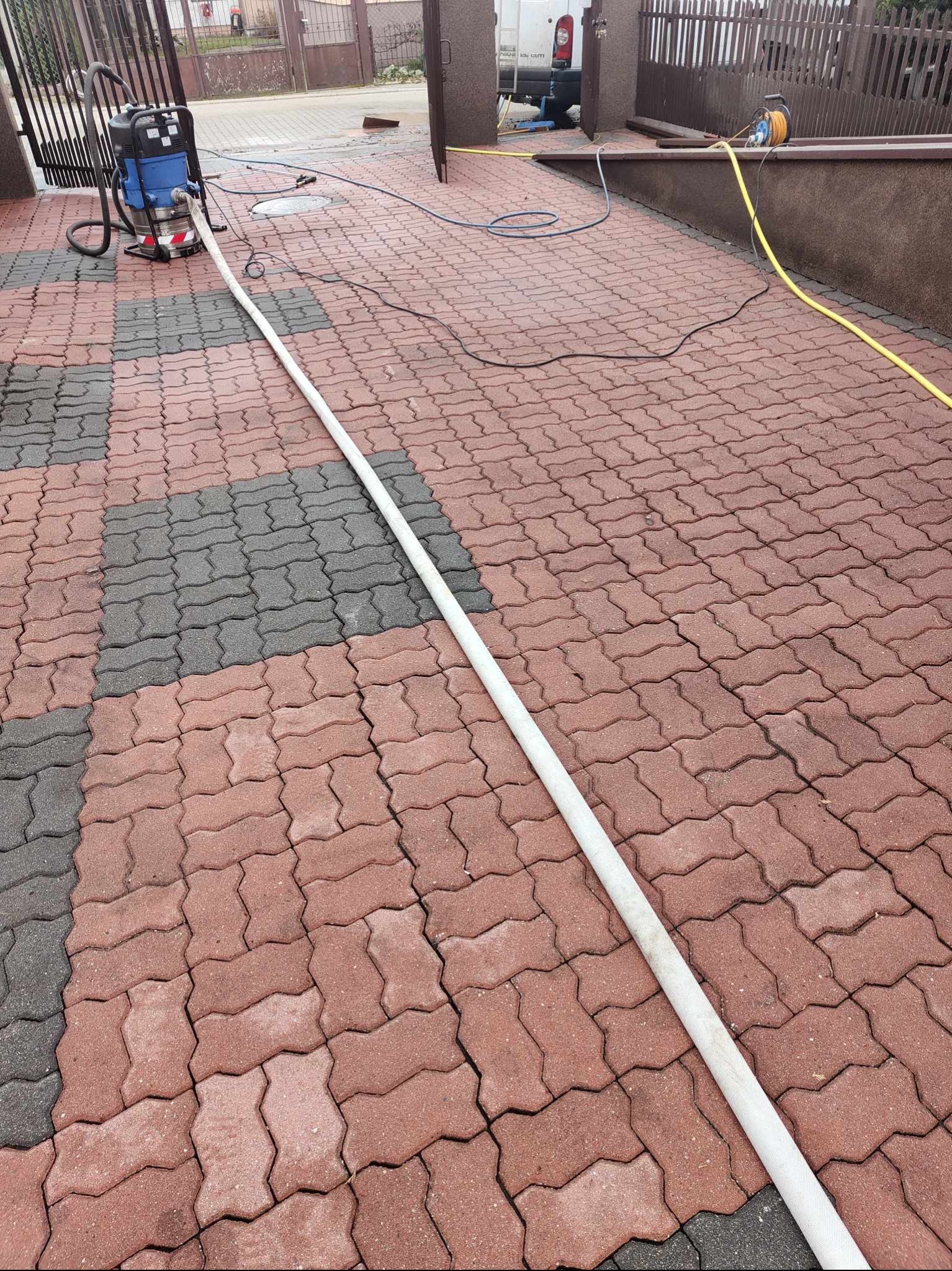 Profesjonalna myjka ciśnieniowa Kärcher podłączona wężem do kostki brukowej w kolorze czerwono-szarym, przygotowana do czyszczenia na posesji przed ogrodzeniem.