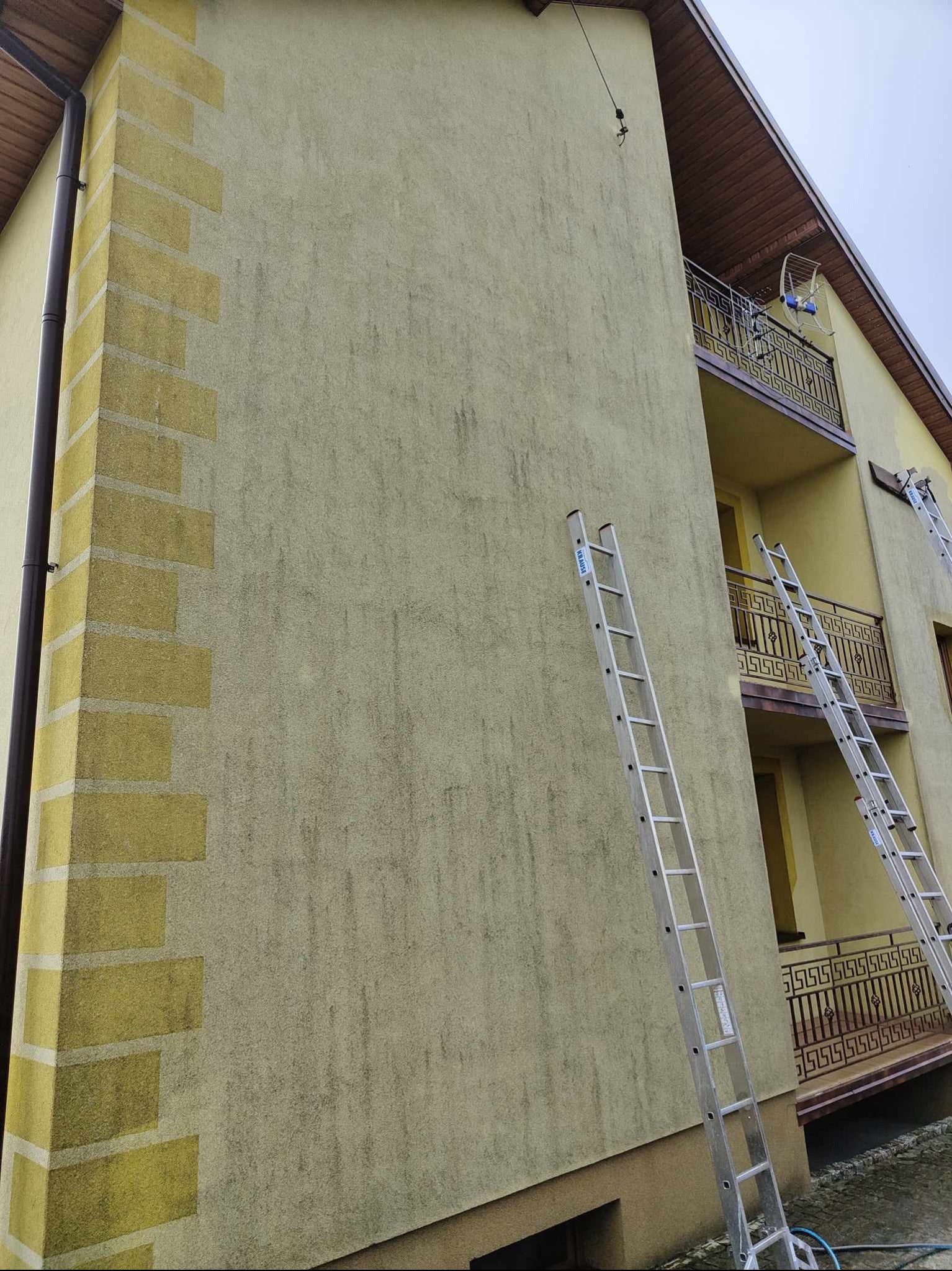 Żółta elewacja domu wielorodzinnego przed malowaniem, widoczne ślady zabrudzeń i dwie aluminiowe drabiny Krause oparte o ścianę.