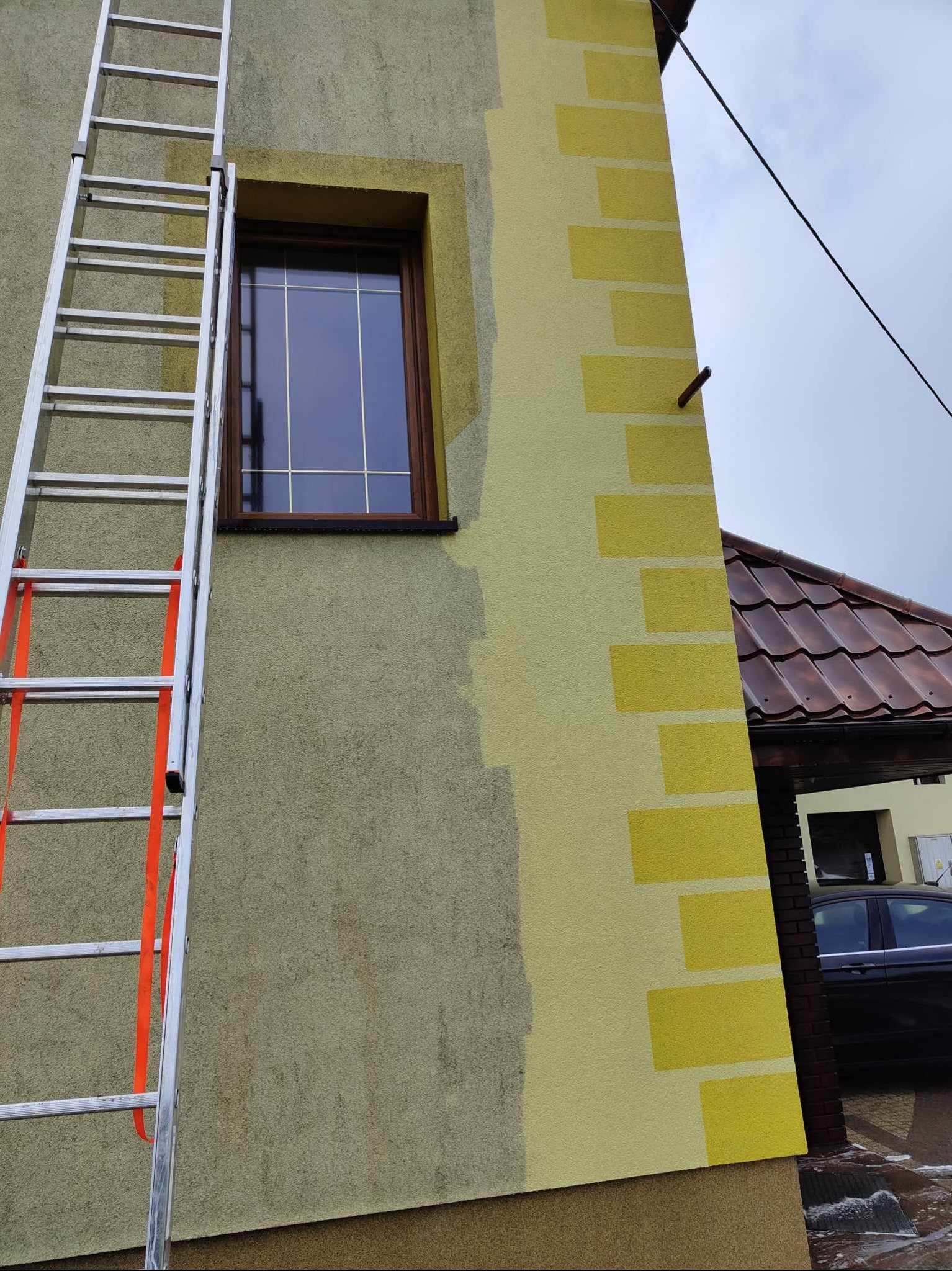 Fragment elewacji budynku z drabina opartą o ścianę, widoczne świeżo nałożone pasy farby w odcieniach żółci, okno z brązową ramą i fragment dachu sąsiedniego budynku.