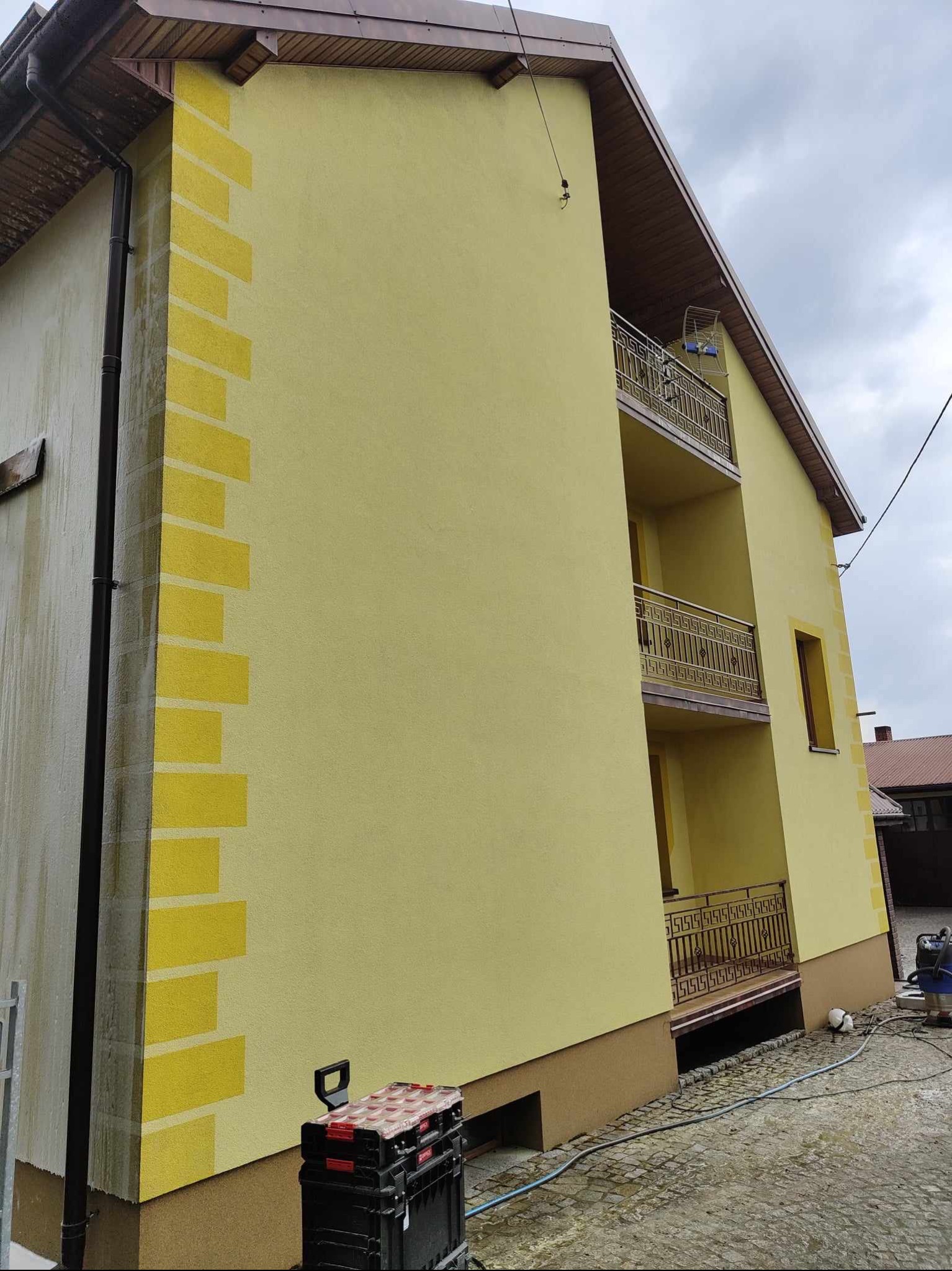 Żółta elewacja domu z dekoracyjnymi elementami w kształcie cegieł na rogu, widoczne balkony z metalowymi balustradami i narzędzia na kostce brukowej.